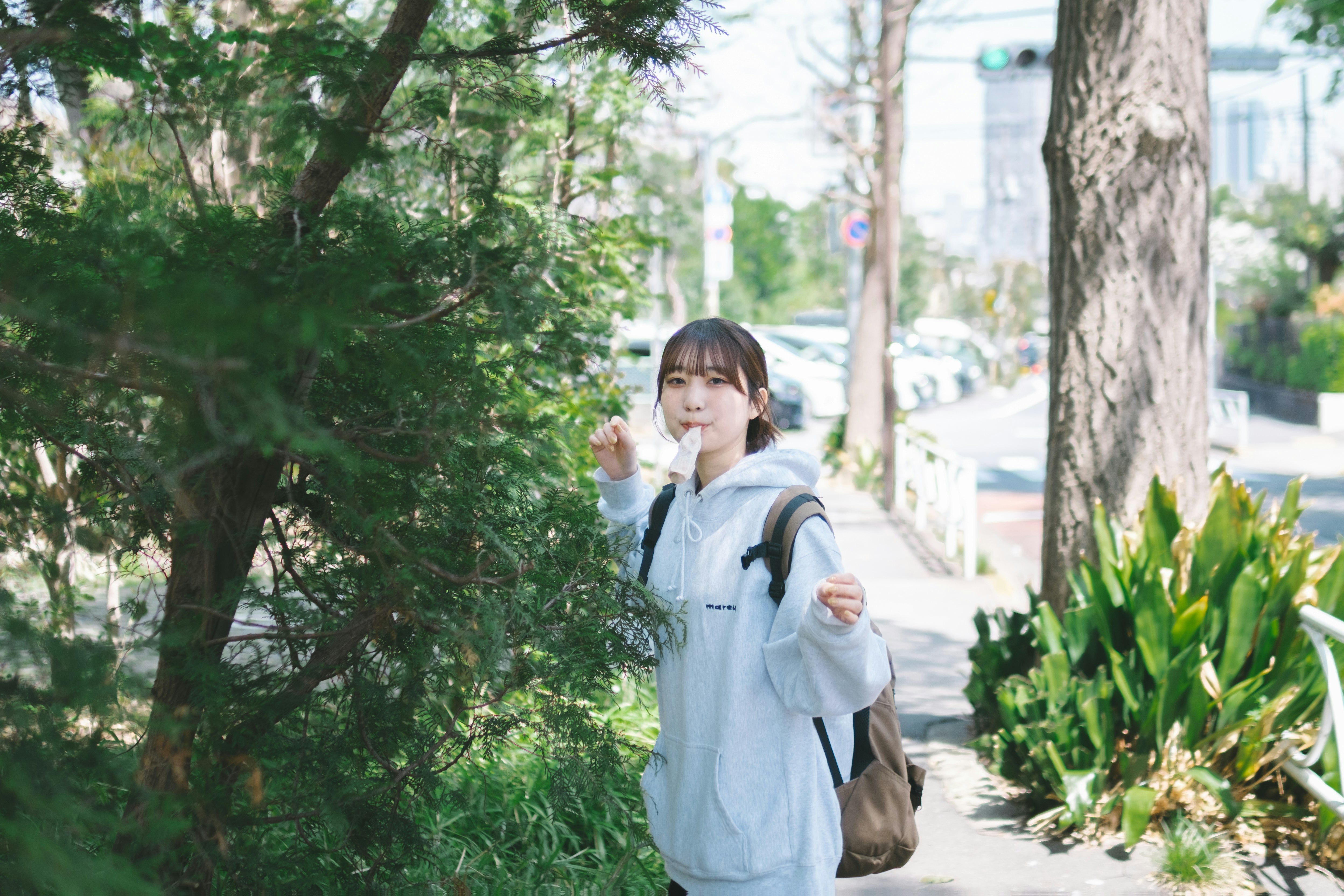 木々の並ぶ道を歩く学生の女性がマスク越しに飲み物を飲む