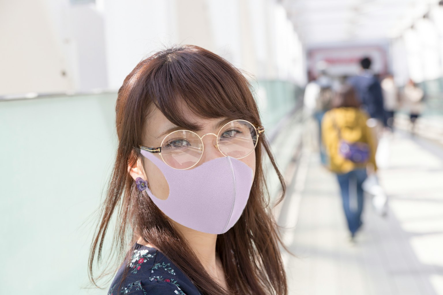 Donna sorridente con maschera viola e occhiali in un corridoio affollato con persone sullo sfondo