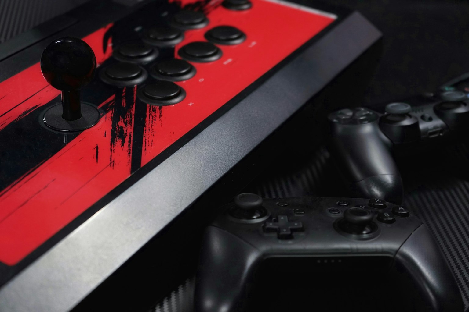 Gậy arcade đỏ với nút đen cạnh hai bộ điều khiển đen trên nền tối