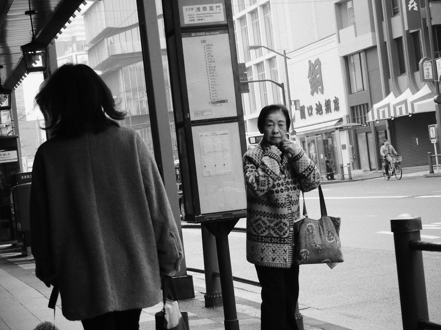 女性が街角で立っているモノクロ写真後ろに別の女性が歩いている