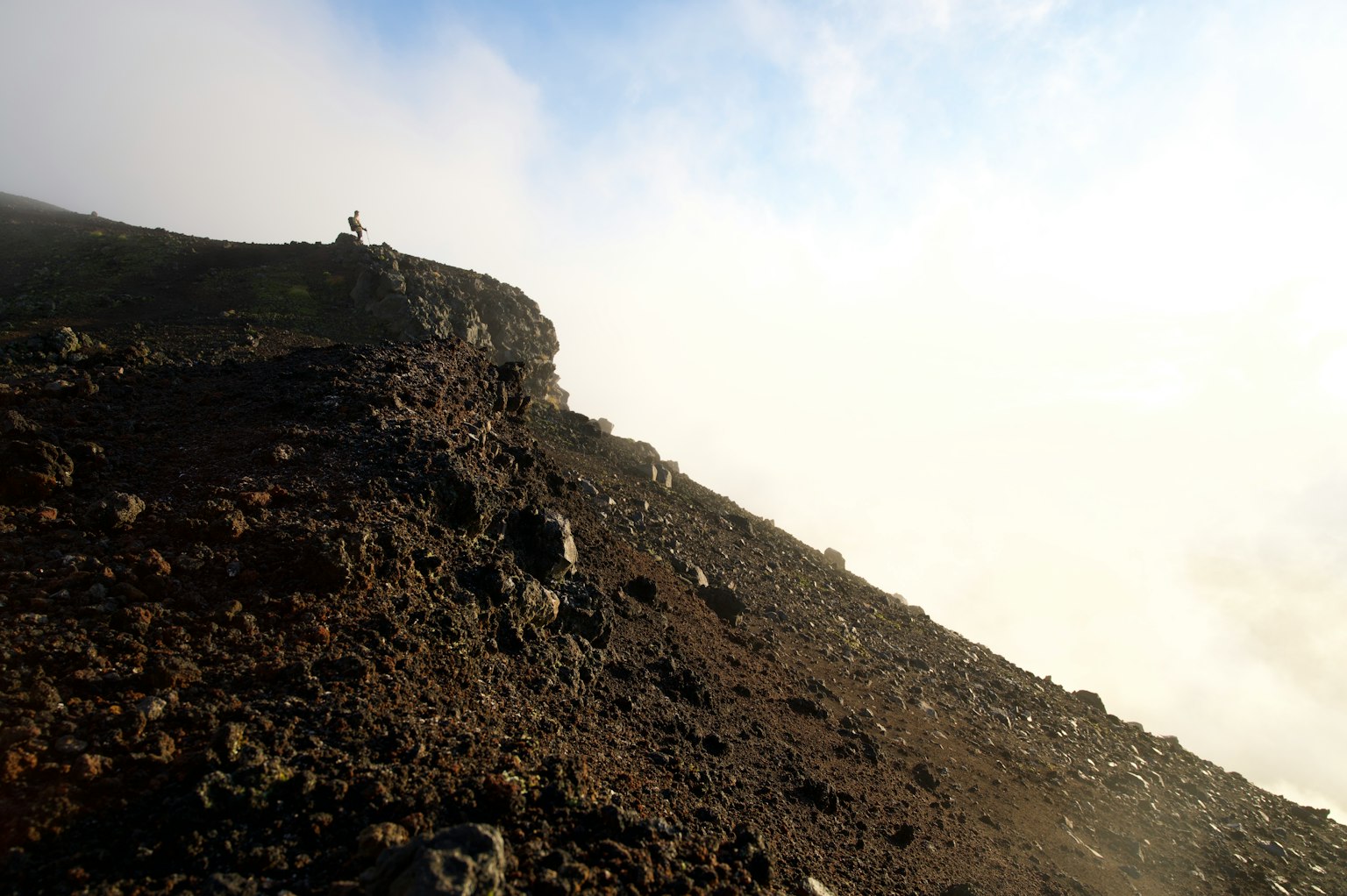 Una persona in piedi su una pendenza vulcanica con nebbia e nuvole sullo sfondo