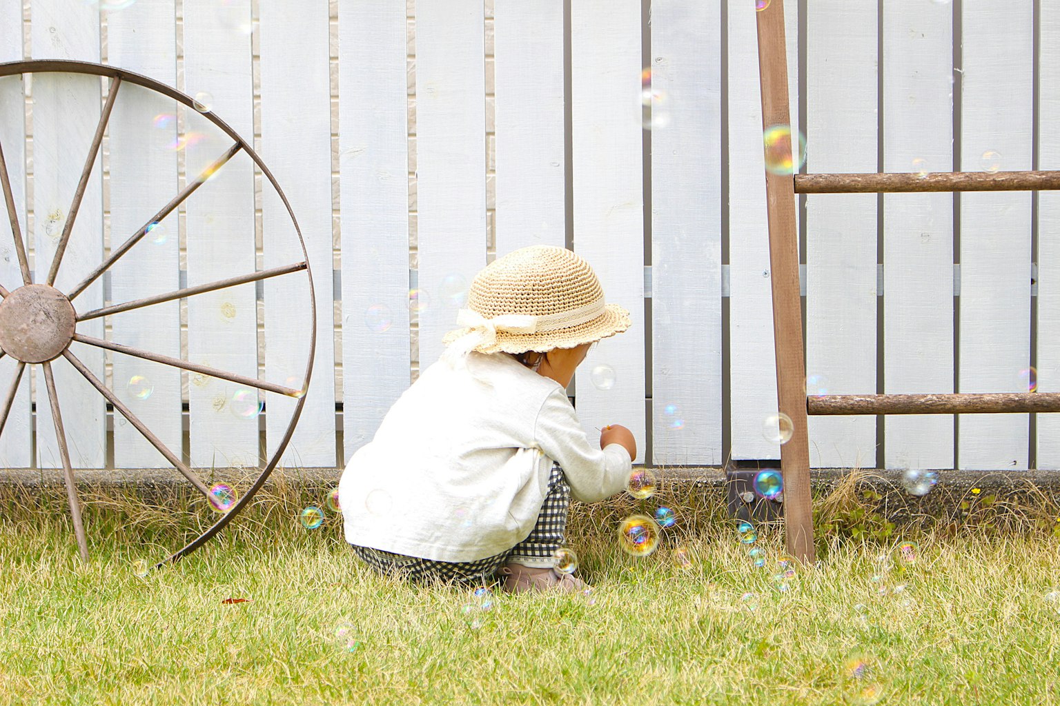 A child discovering bubbles on the grass with a white fence and an old wheel in the background