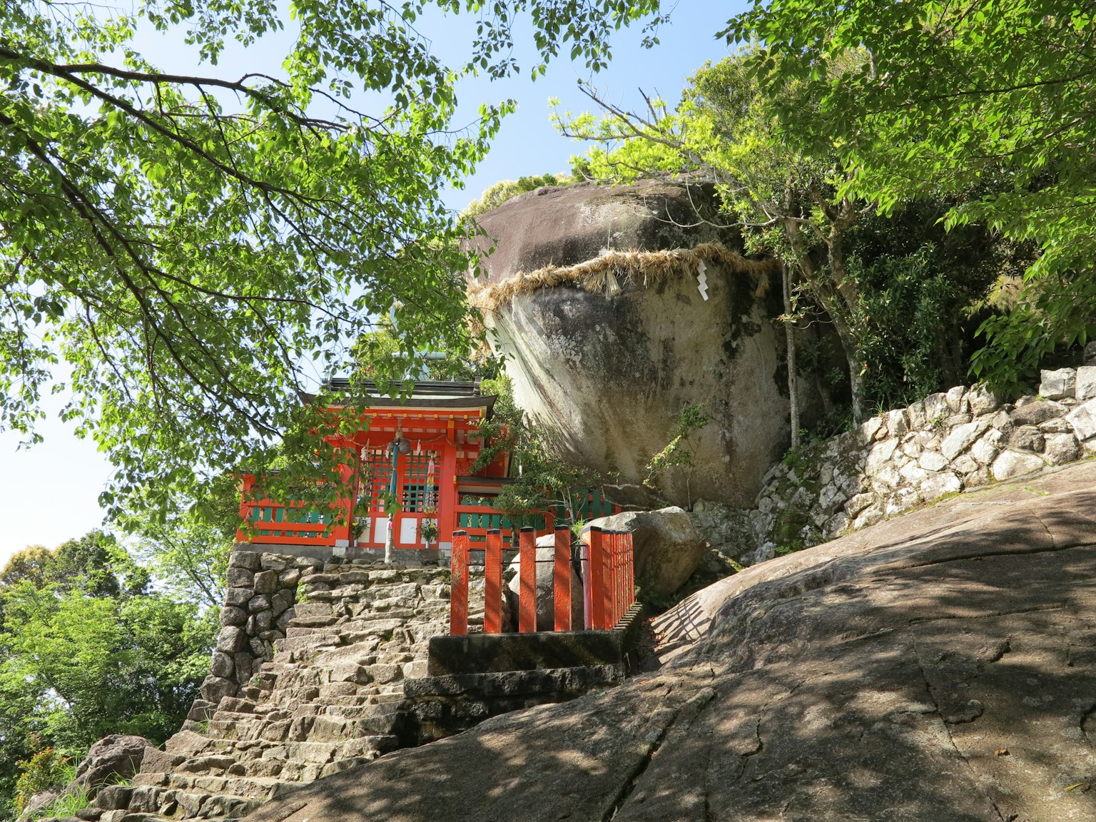 Pemandangan kuil dengan gerbang torii merah dan batu besar dikelilingi pohon hijau