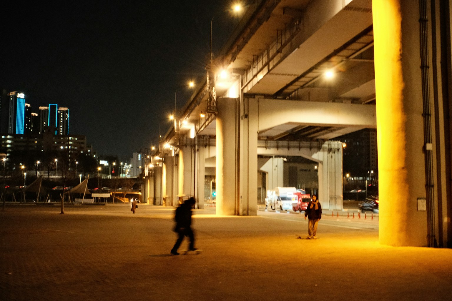 Nächtliche Stadtlandschaft mit Menschen unter einer Autobahn, beleuchtet von Straßenlaternen und Gebäuden