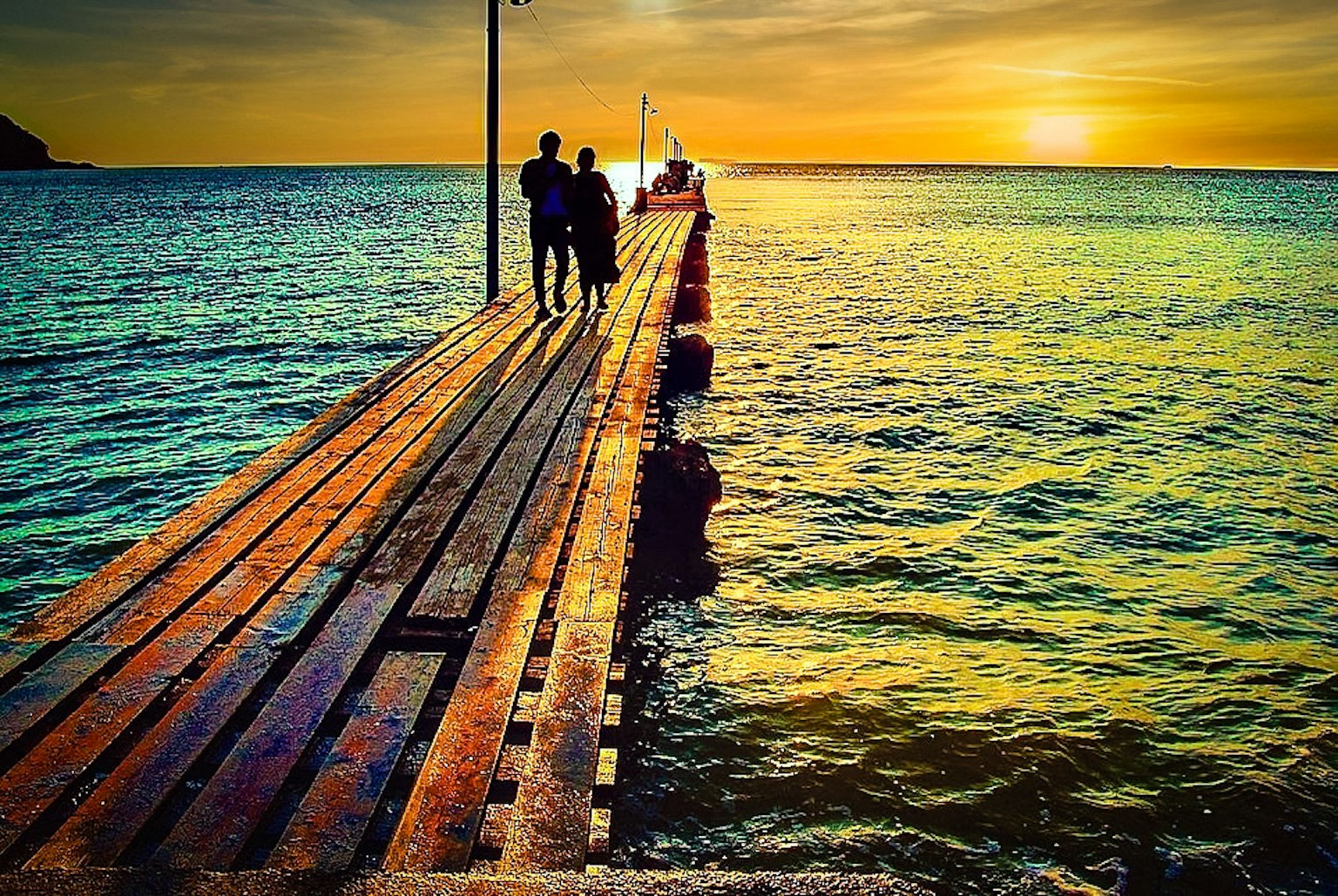 Pareja caminando por un muelle de madera hacia el atardecer sobre el mar