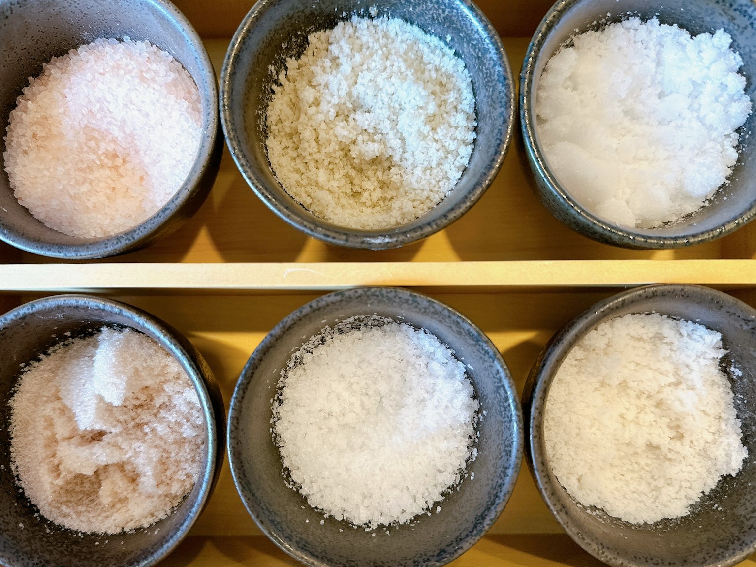 Seis tazones grises llenos de diferentes tipos de sal dispuestos en una bandeja de madera