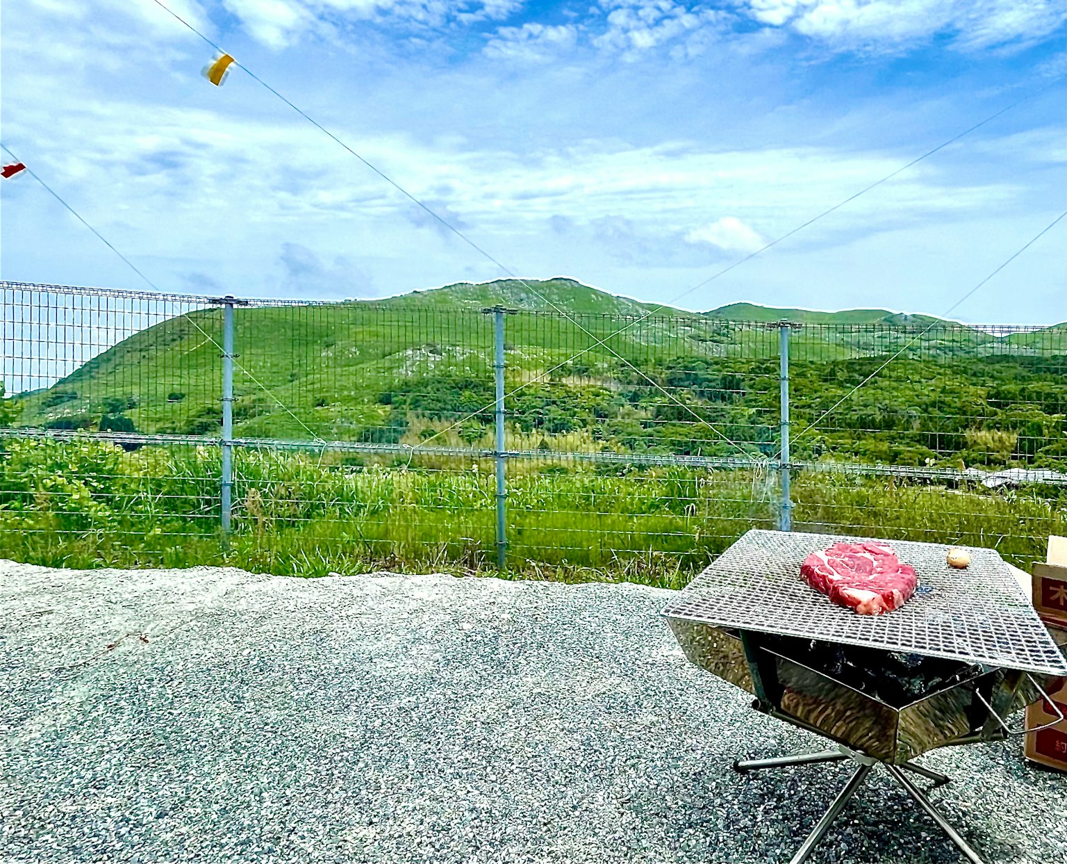 Grilltisch mit Fleisch vor einer Kulisse aus grünen Hügeln und blauem Himmel