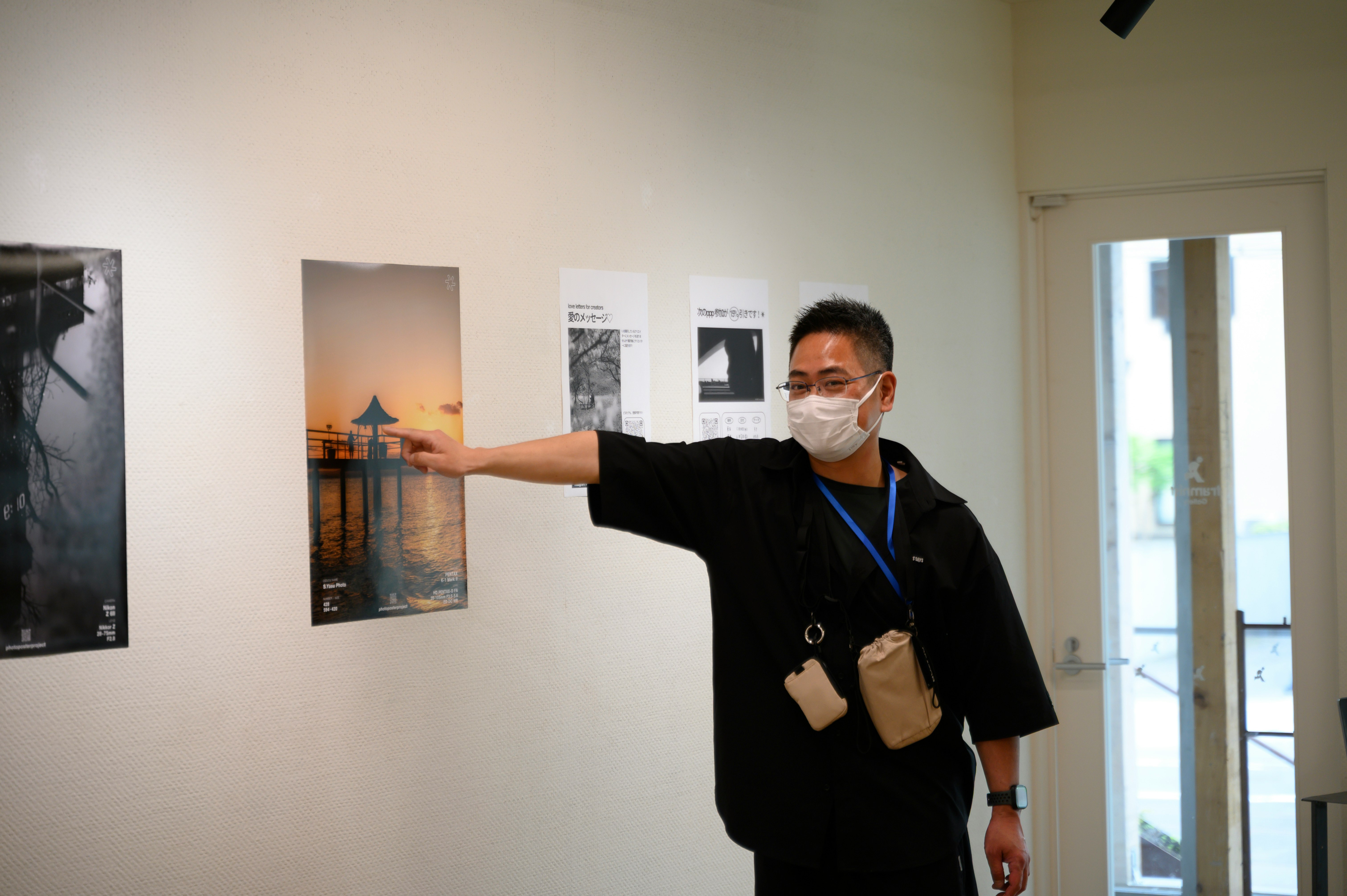 展示ポスターを指さすマスク姿の案内スタッフのいる室内