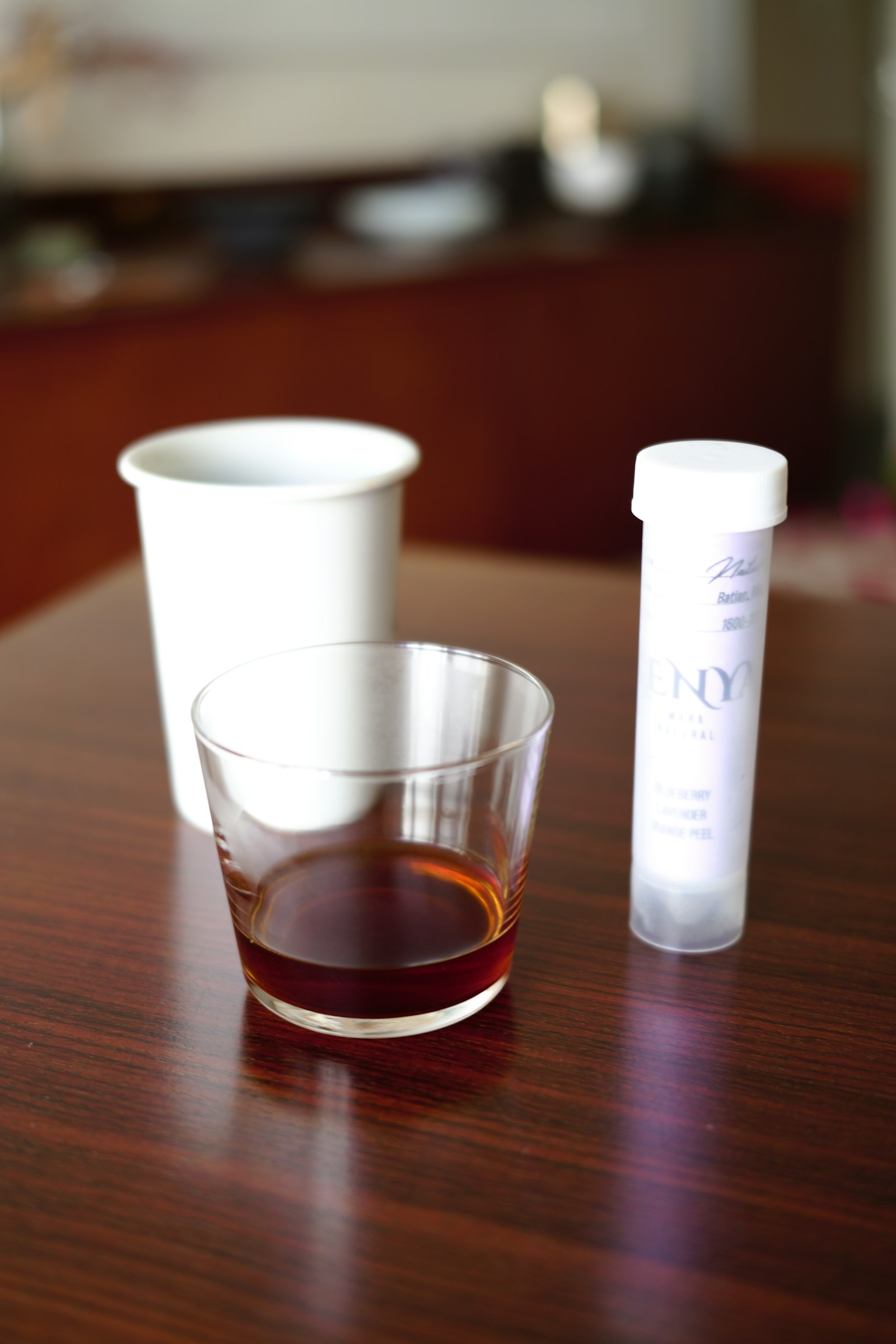 A clear glass with a brown beverage and a white plastic container on a wooden table