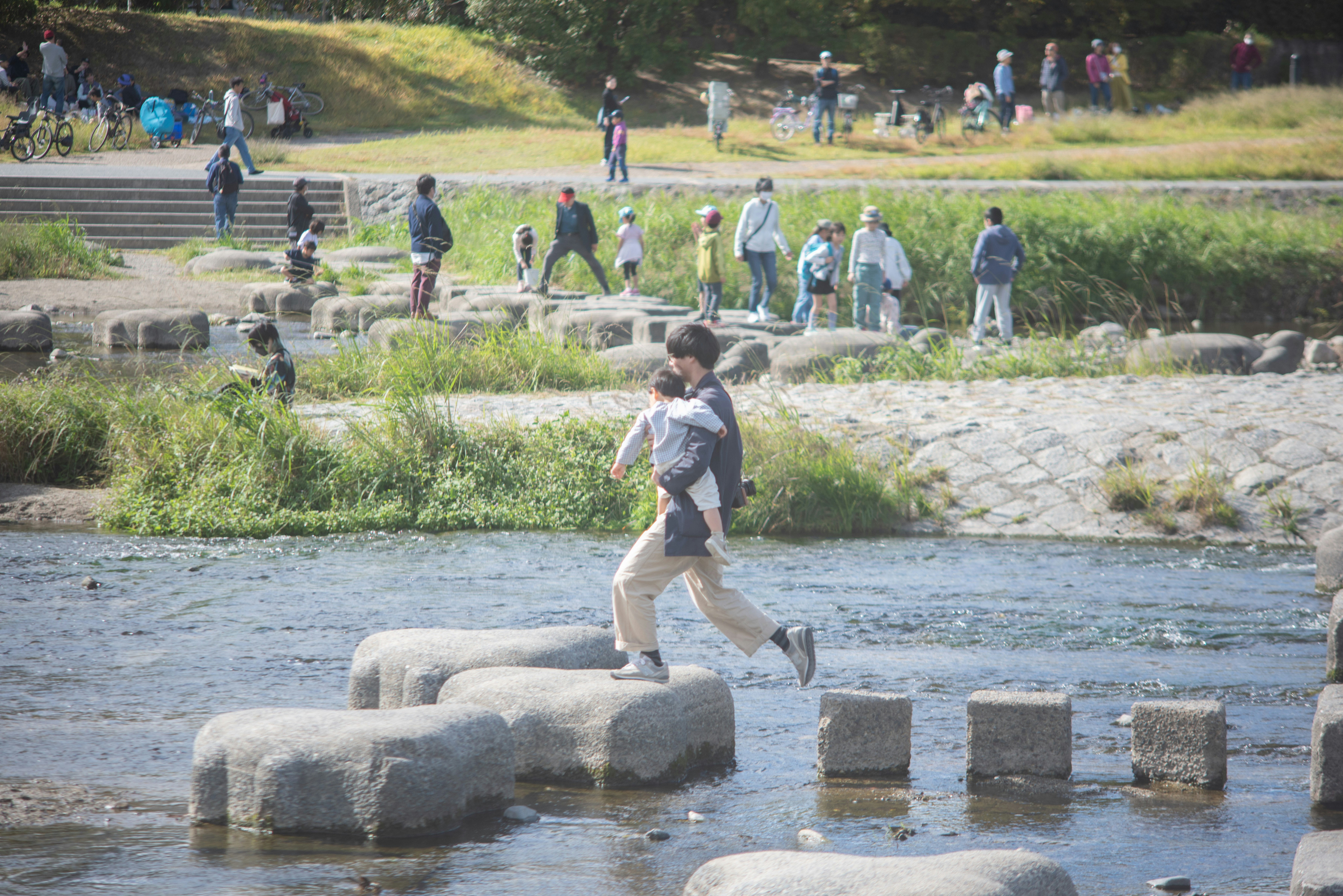 川の石を渡る子供たちと周囲の人々がいる公園の風景