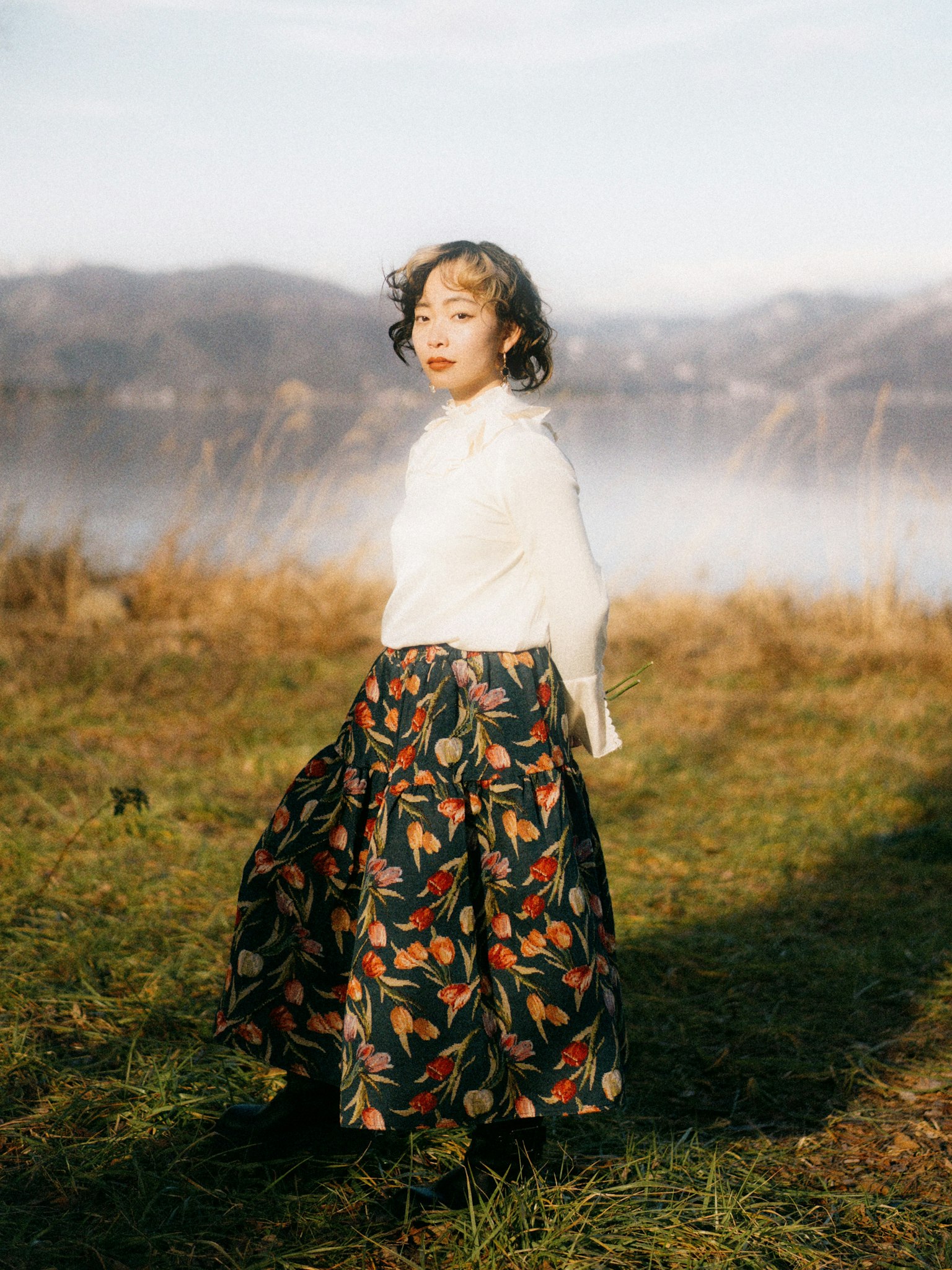 A woman standing near a lake wearing a white blouse and floral skirt