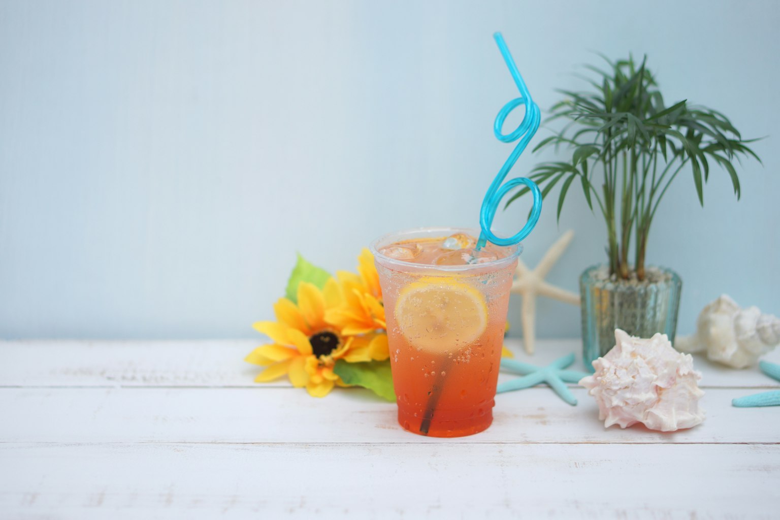 Đồ uống màu cam với lát chanh và ống hút màu xanh bên cạnh hoa hướng dương và cây trên nền xanh nhạt