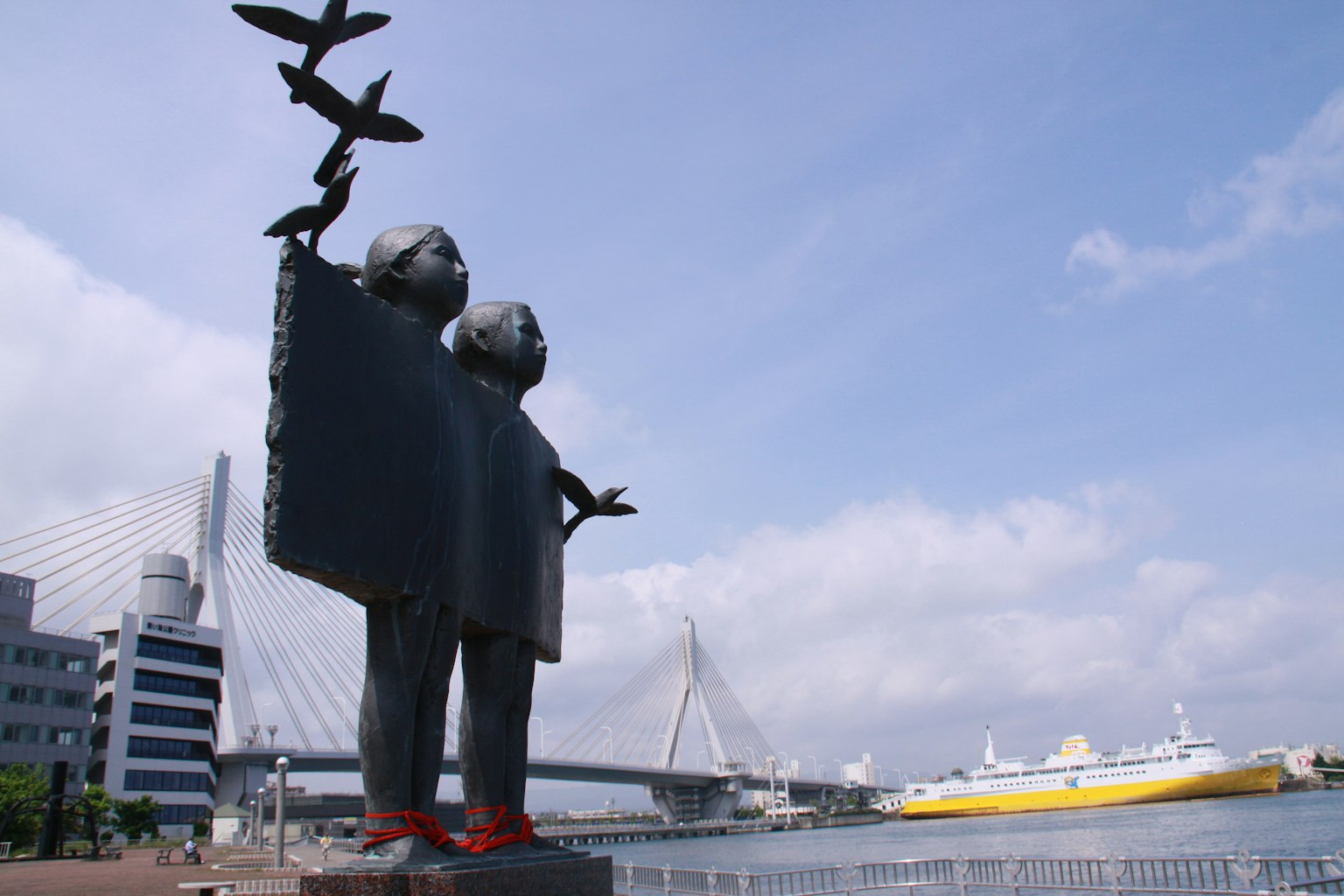 Skulptur von zwei Kindern, die fliegende Vögel halten, mit einer Brücke und einem Schiff im Hintergrund