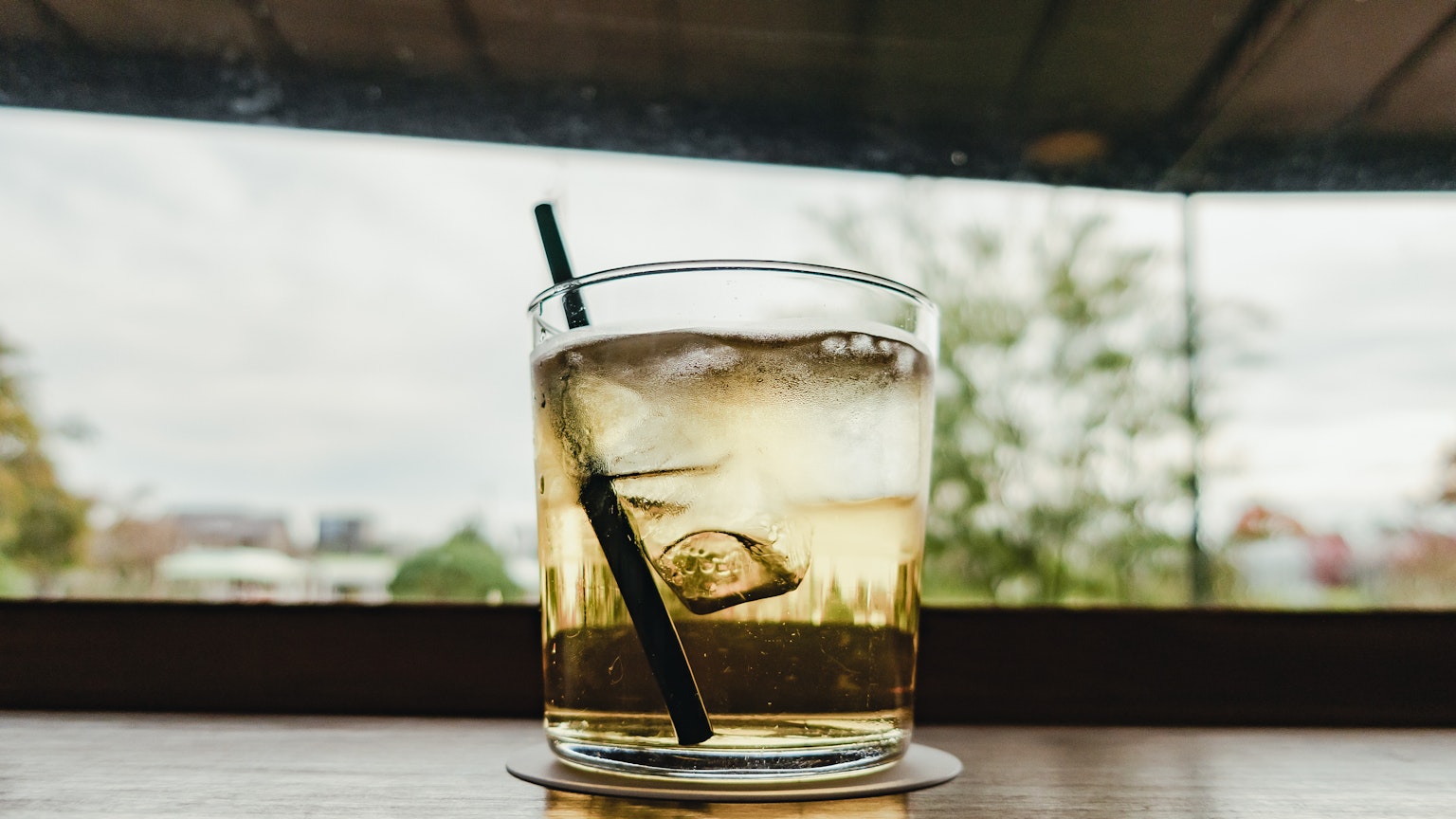 Bicchiere di whisky con ghiaccio e una cannuccia nera con sfondo di alberi verdi visibili attraverso una finestra