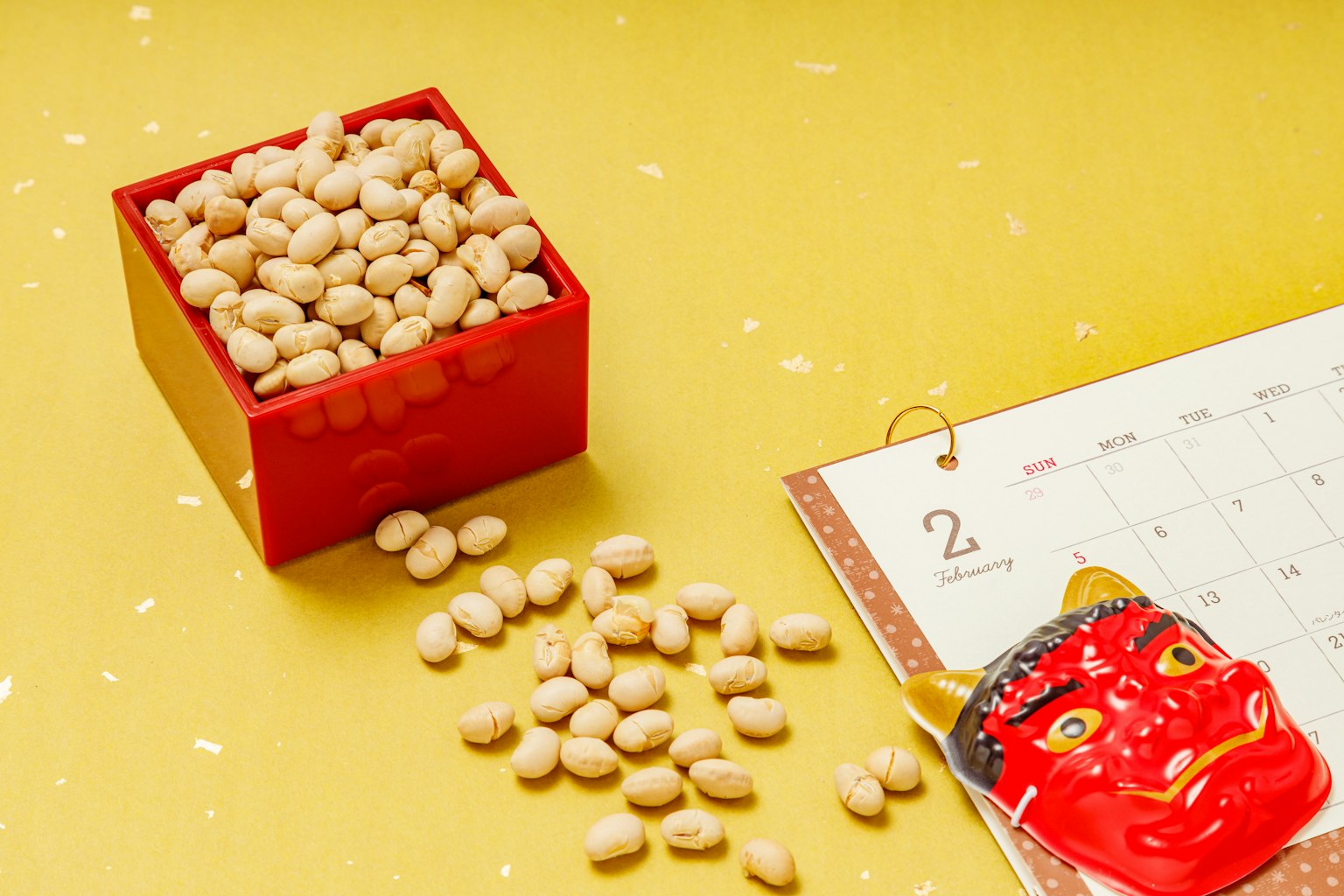 Image of a red box filled with beans and scattered beans alongside a demon mask on a yellow background