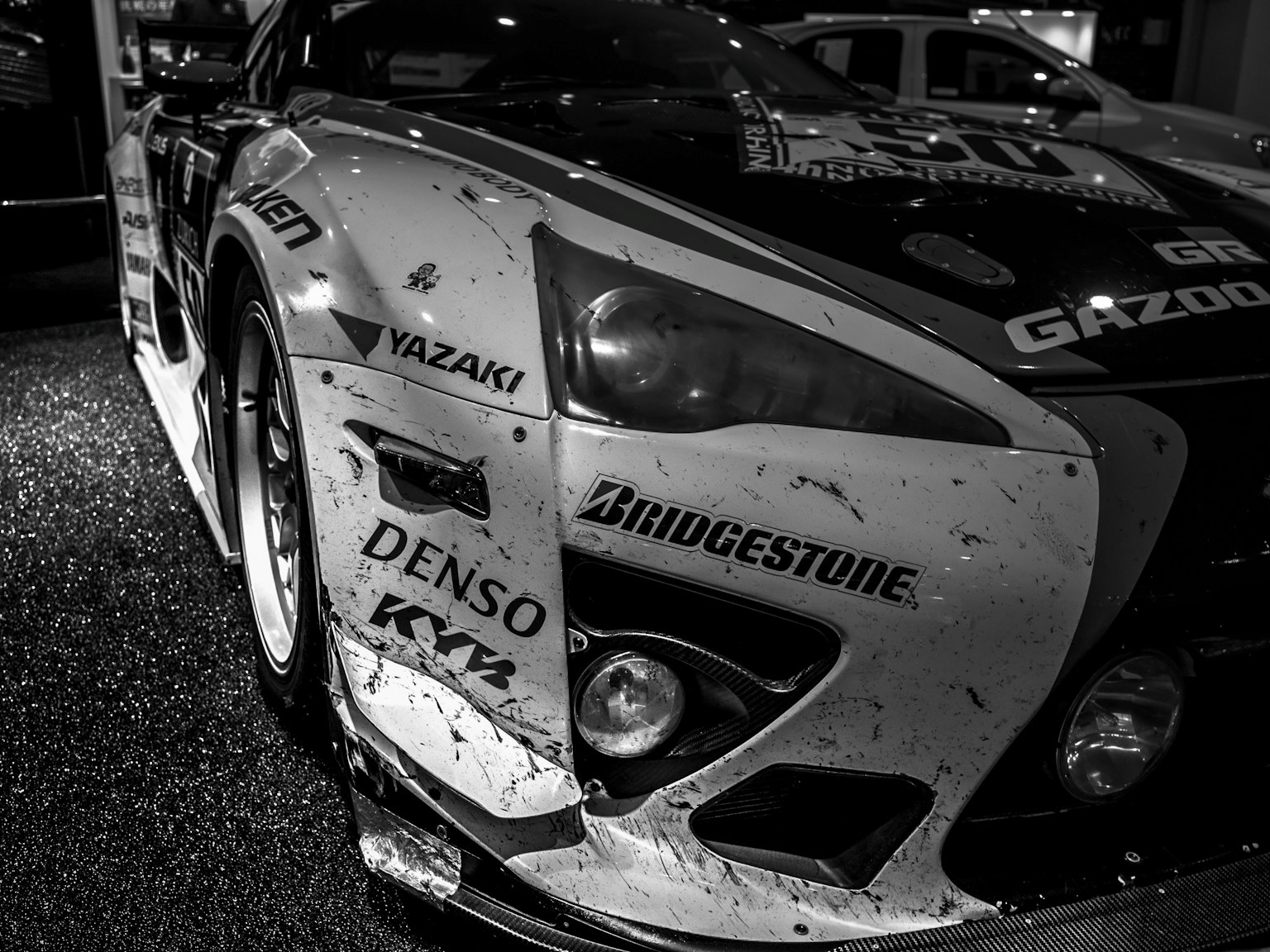 Kedekatan mobil balap hitam dan putih dengan logo Bridgestone menunjukkan goresan dan kotoran