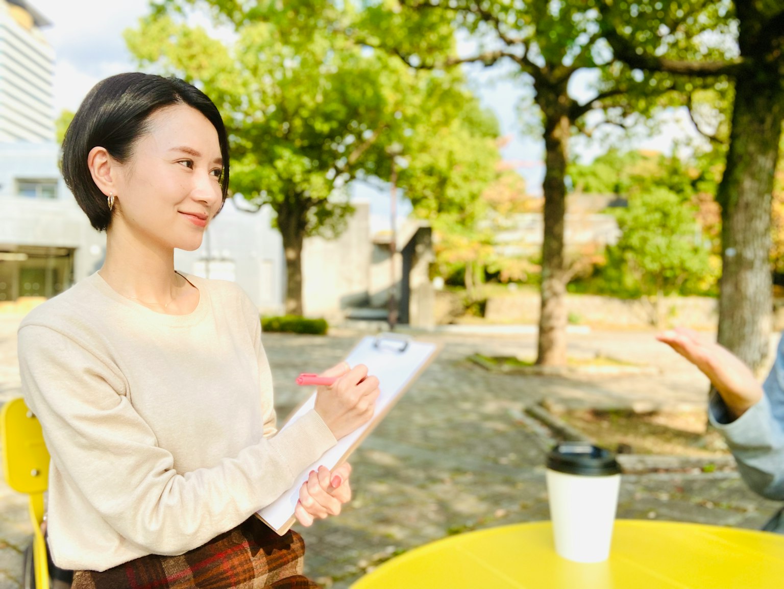 Femme interviewée dans un parc tenant un clipboard à une table jaune avec des arbres verts en arrière-plan