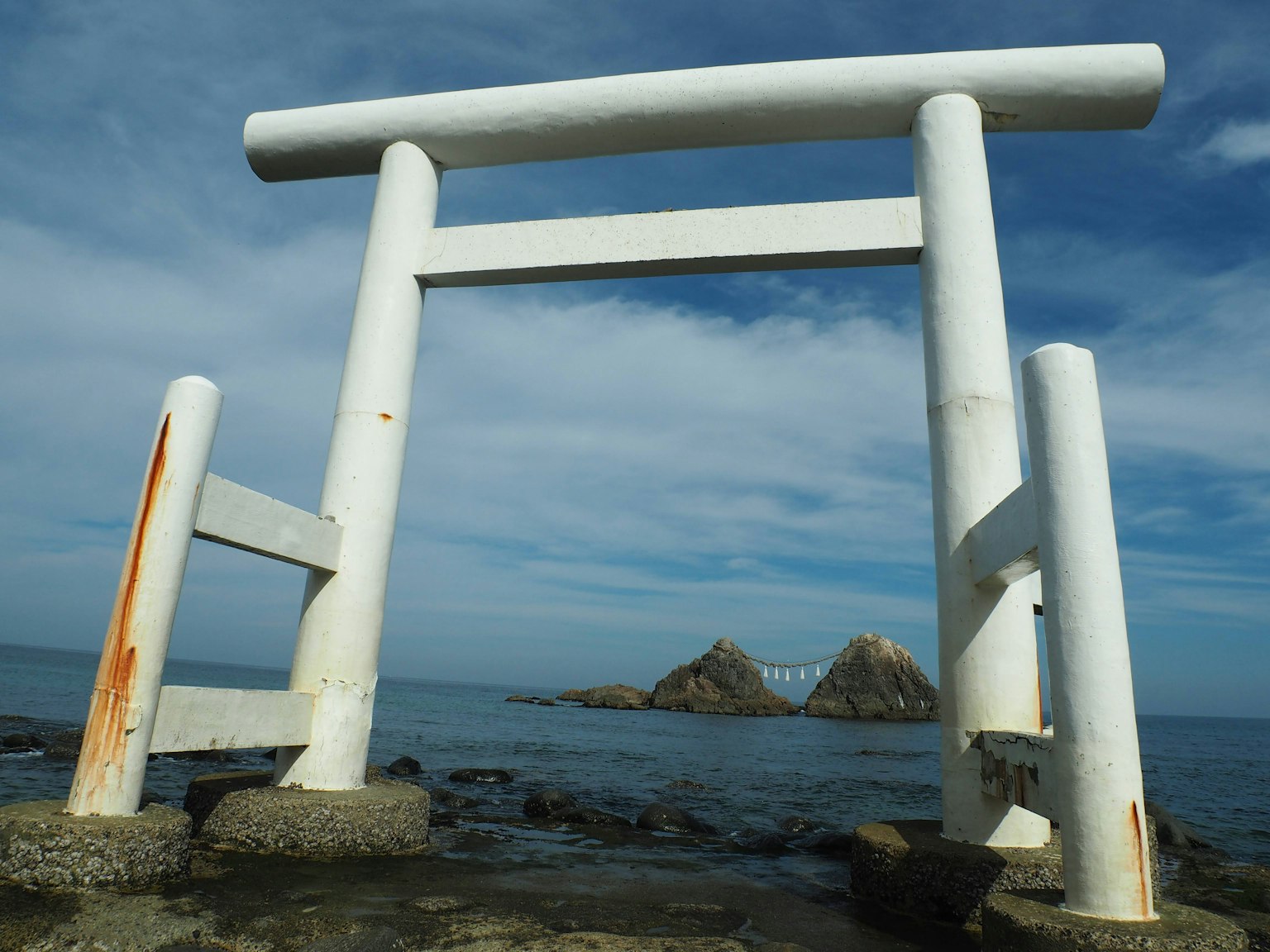 Gerbang torii putih berdiri di dekat laut dengan batu-batu di latar belakang