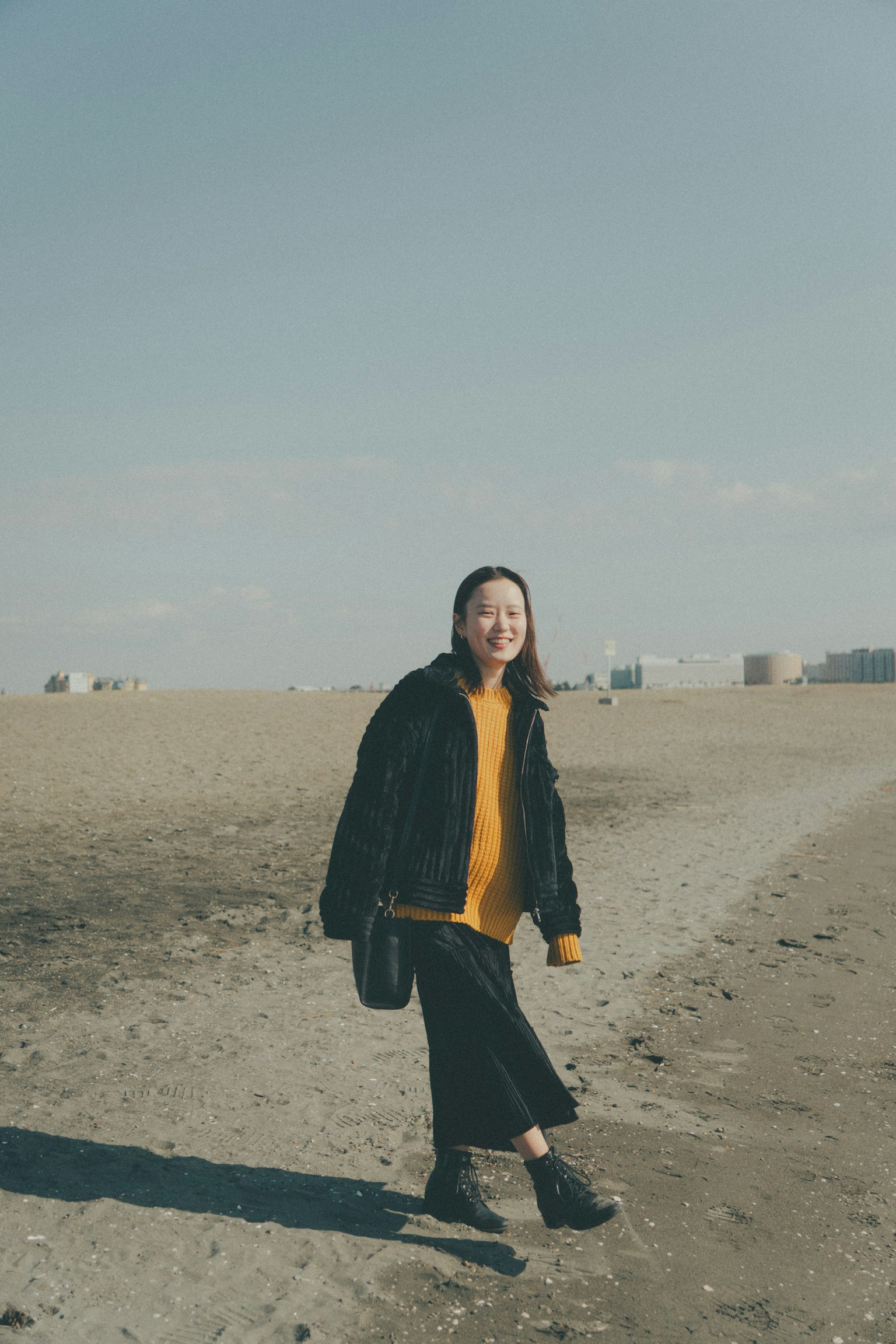 Una mujer caminando por la playa con una chaqueta negra y un suéter amarillo