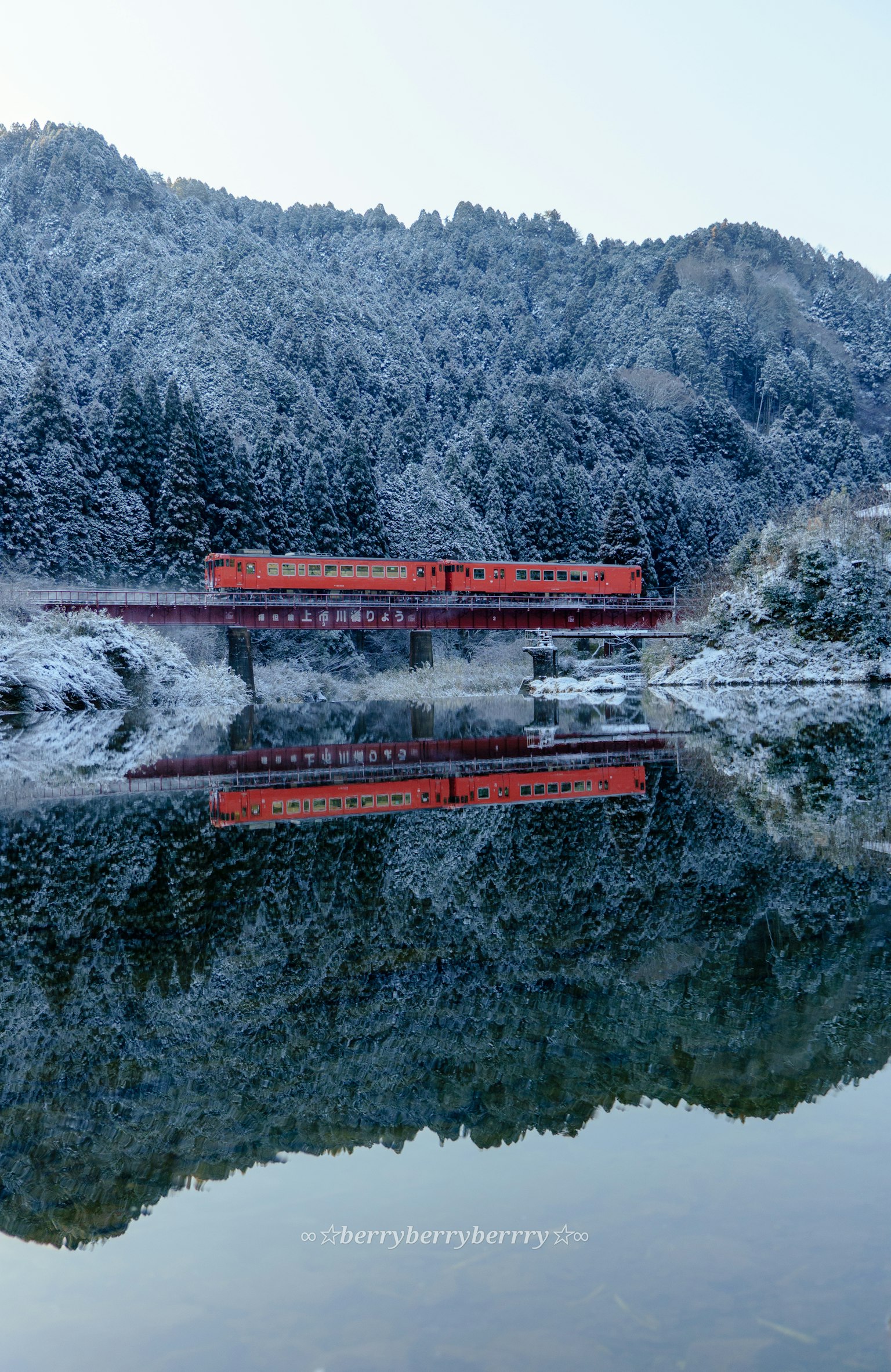 รถไฟสีแดงข้ามสะพานที่มีหิมะกับการสะท้อนในน้ำ