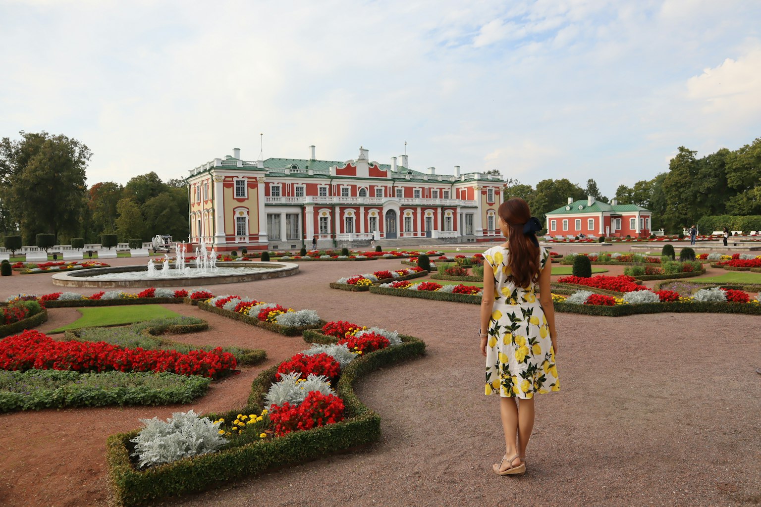 Donna in abito floreale che cammina in un giardino con un edificio storico sullo sfondo