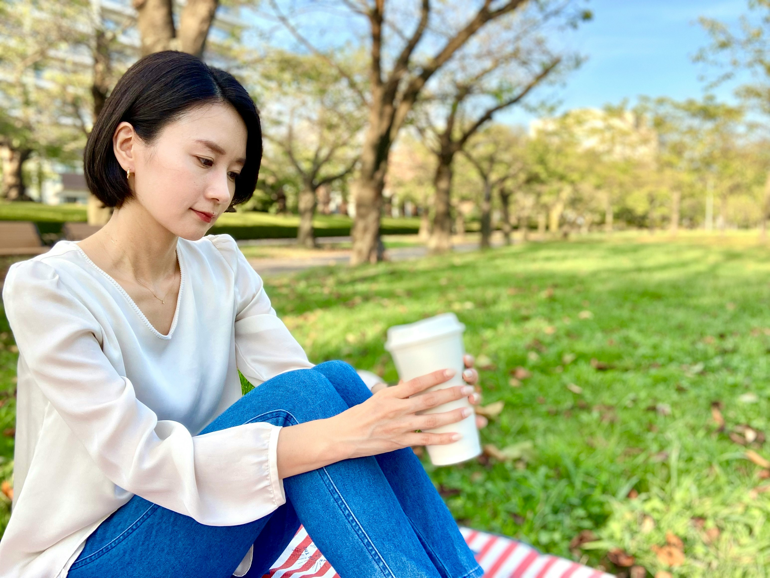 người phụ nữ ngồi trên bãi cỏ công viên cầm cốc cà phê