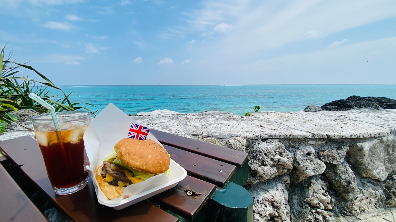Ein Burger und ein Eistee auf einem Holztisch mit blauem Ozean im Hintergrund