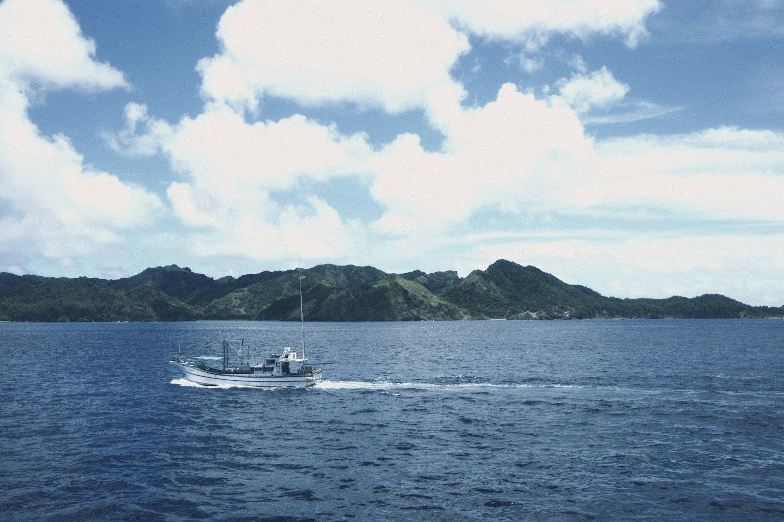 一艘小船在藍色海水上航行，白雲下有綠色山丘