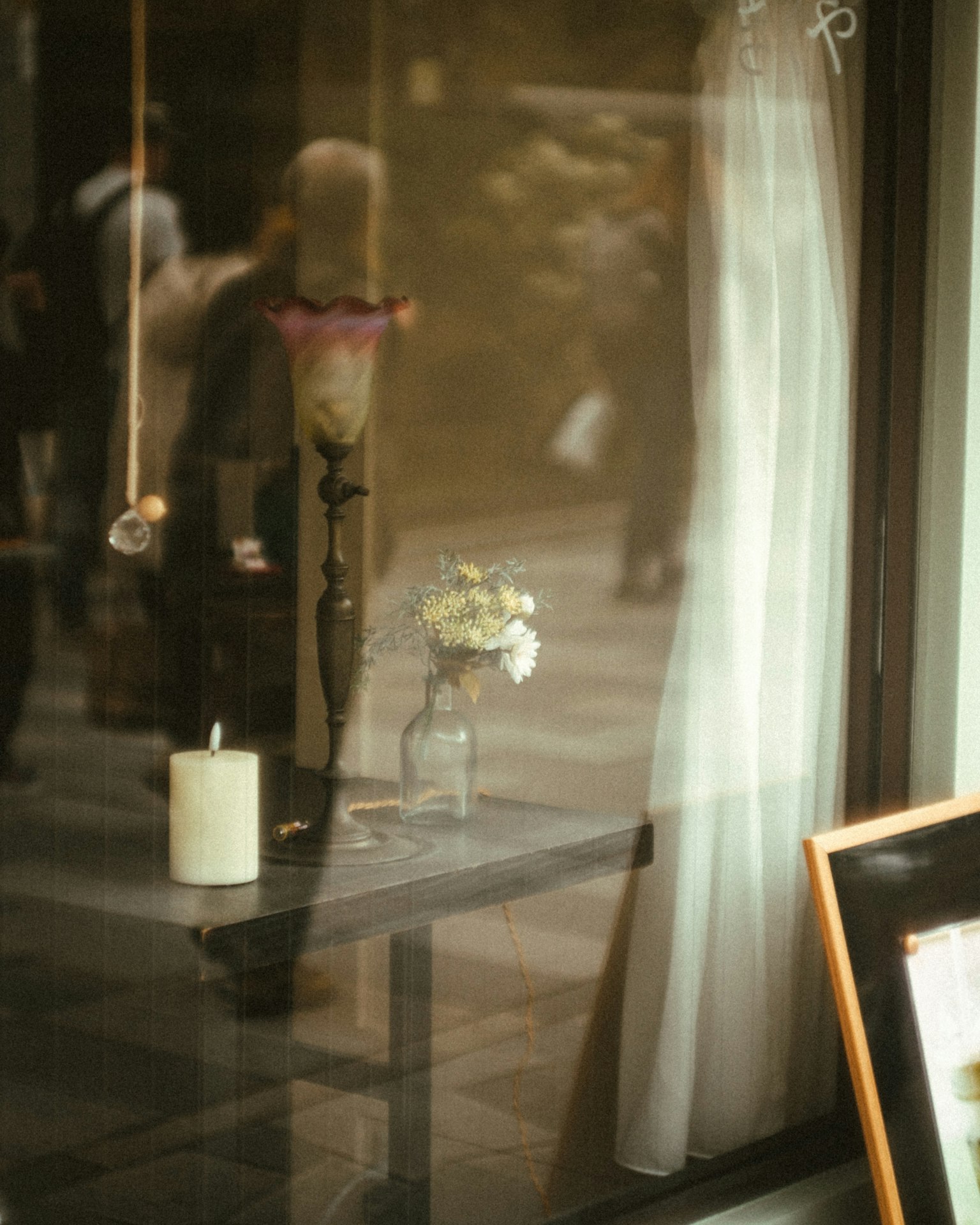 A cozy interior scene with a vase of flowers and a candle by the window reflections of passersby visible