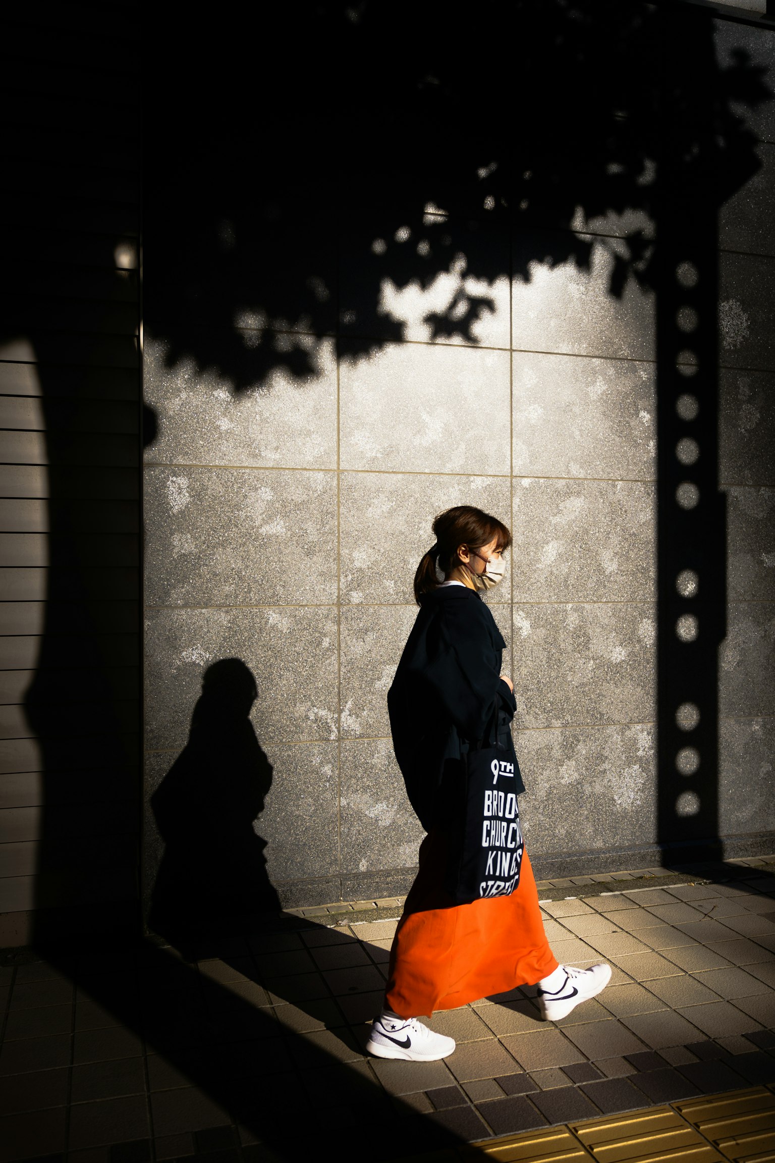 穿著黑色外套和橙色裙子的女性在城市街道上走，伴隨著她的影子