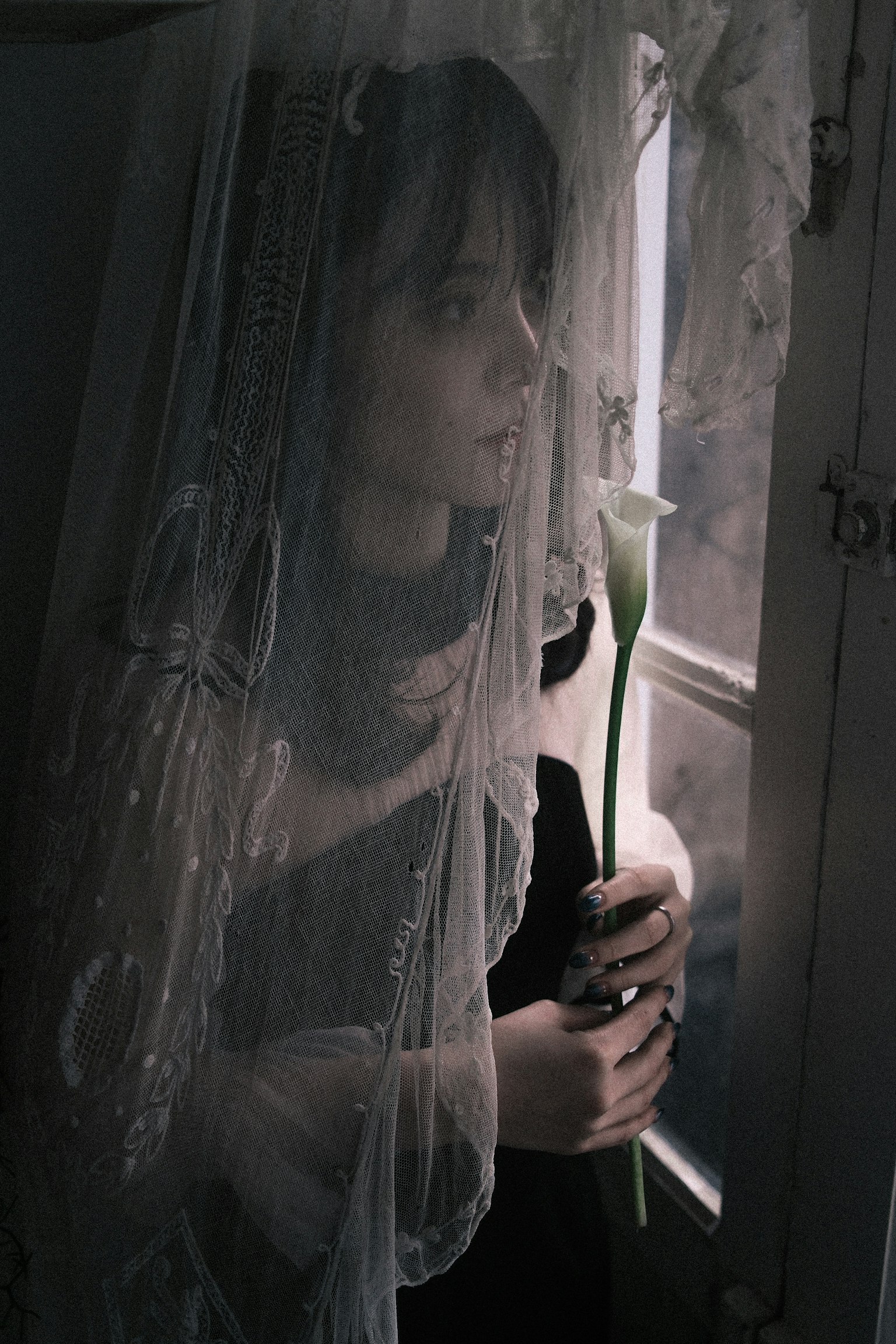 窓のそばに立つ女性が白い花を持っている薄暗い部屋の中でベールをかぶっている