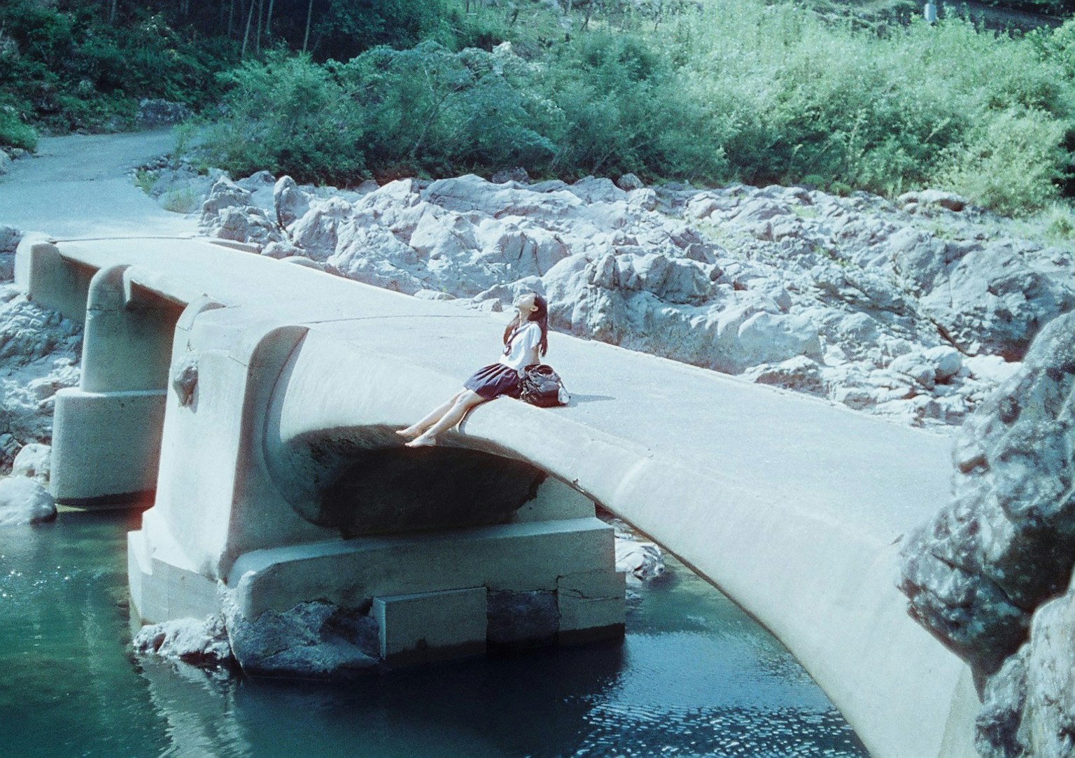 Frau sitzt auf einer Betonbrücke umgeben von Grün in der Nähe von fließendem Wasser und Felsen