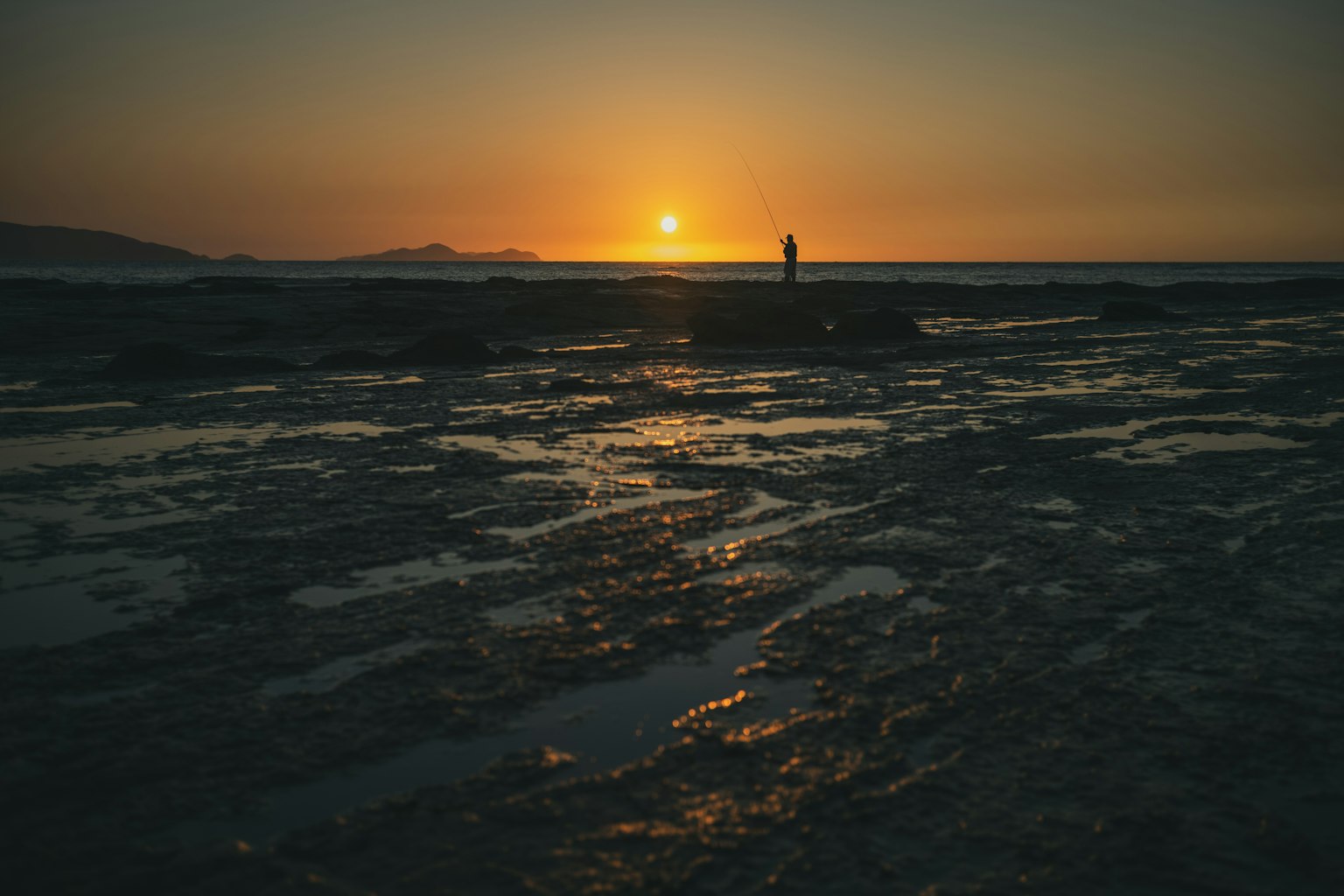 Une personne se tenant sur une côte rocheuse au coucher du soleil avec des reflets sur l'eau