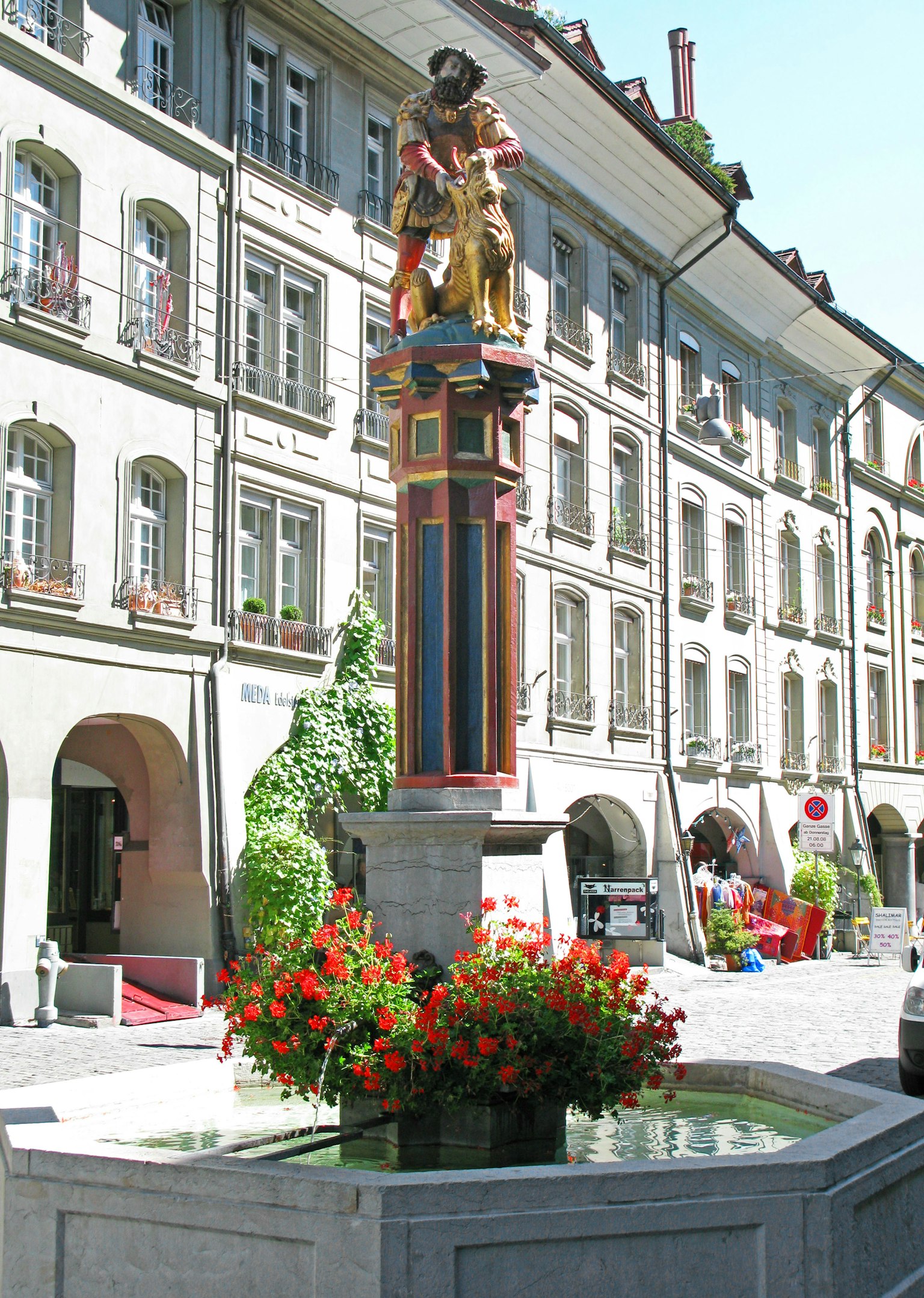 Statue colorée se tenant dans une place avec une fontaine et des fleurs en fleurs