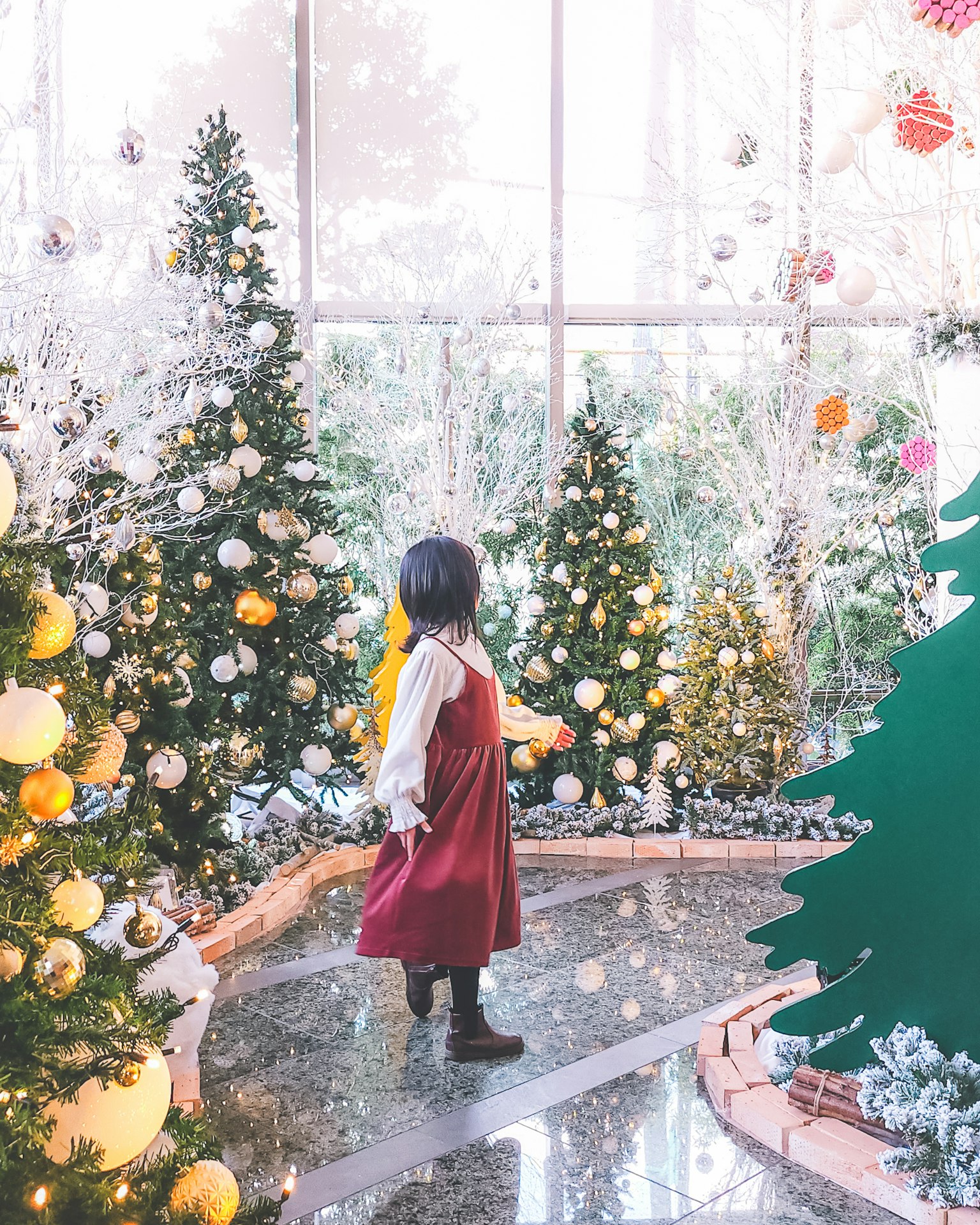 Anak dengan gaun merah dikelilingi oleh pohon Natal yang dihias hiasan cerah dan suasana meriah