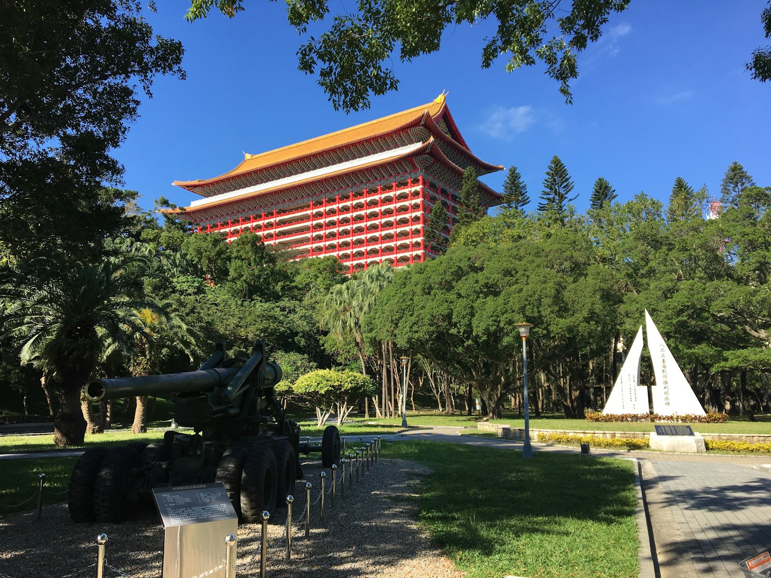 View of a red building surrounded by green trees with a white pyramid sculpture in a park