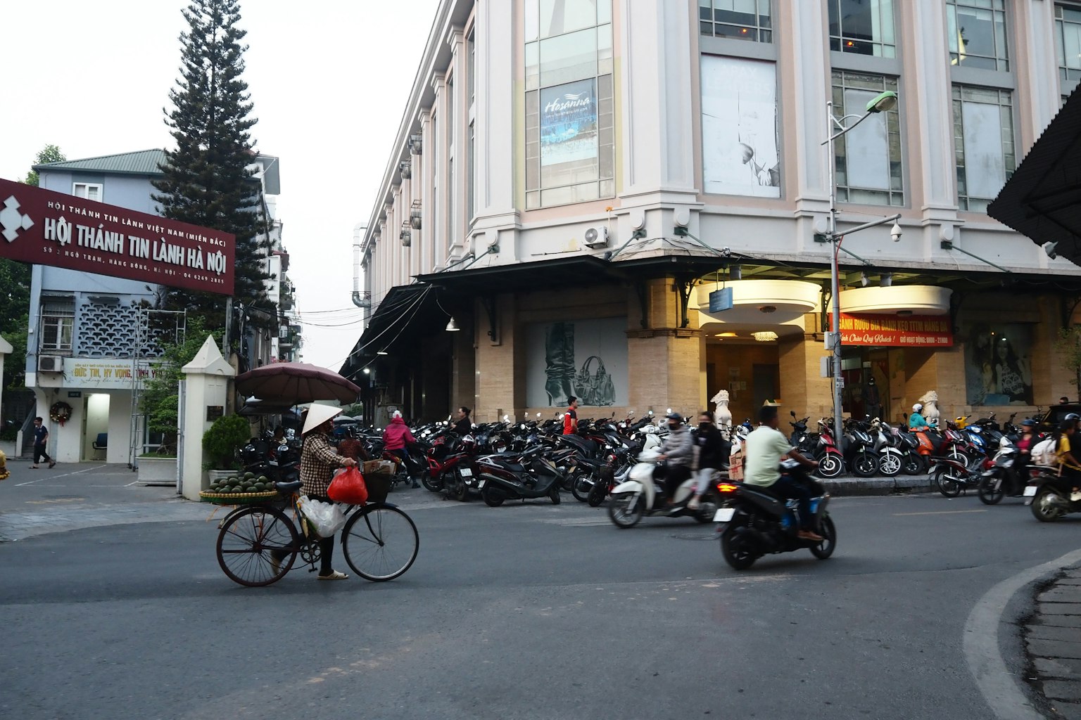 繁忙的交叉口有摩托車和自行車 背景中有商業建築 許多停放的摩托車