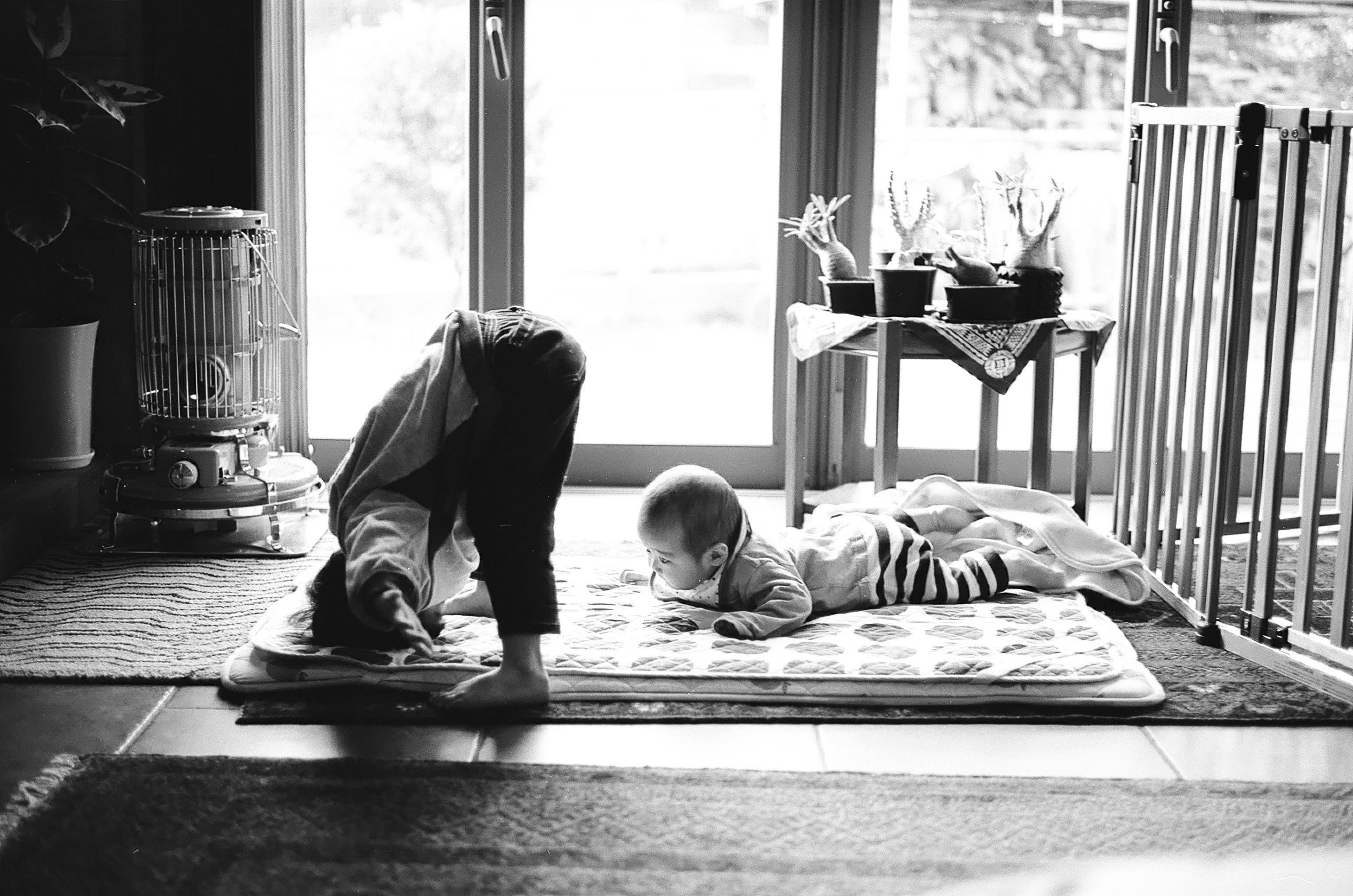 Cảnh trong nhà: em bé chơi trên tấm thảm gần cửa sổ, cạnh đó có người lớn
