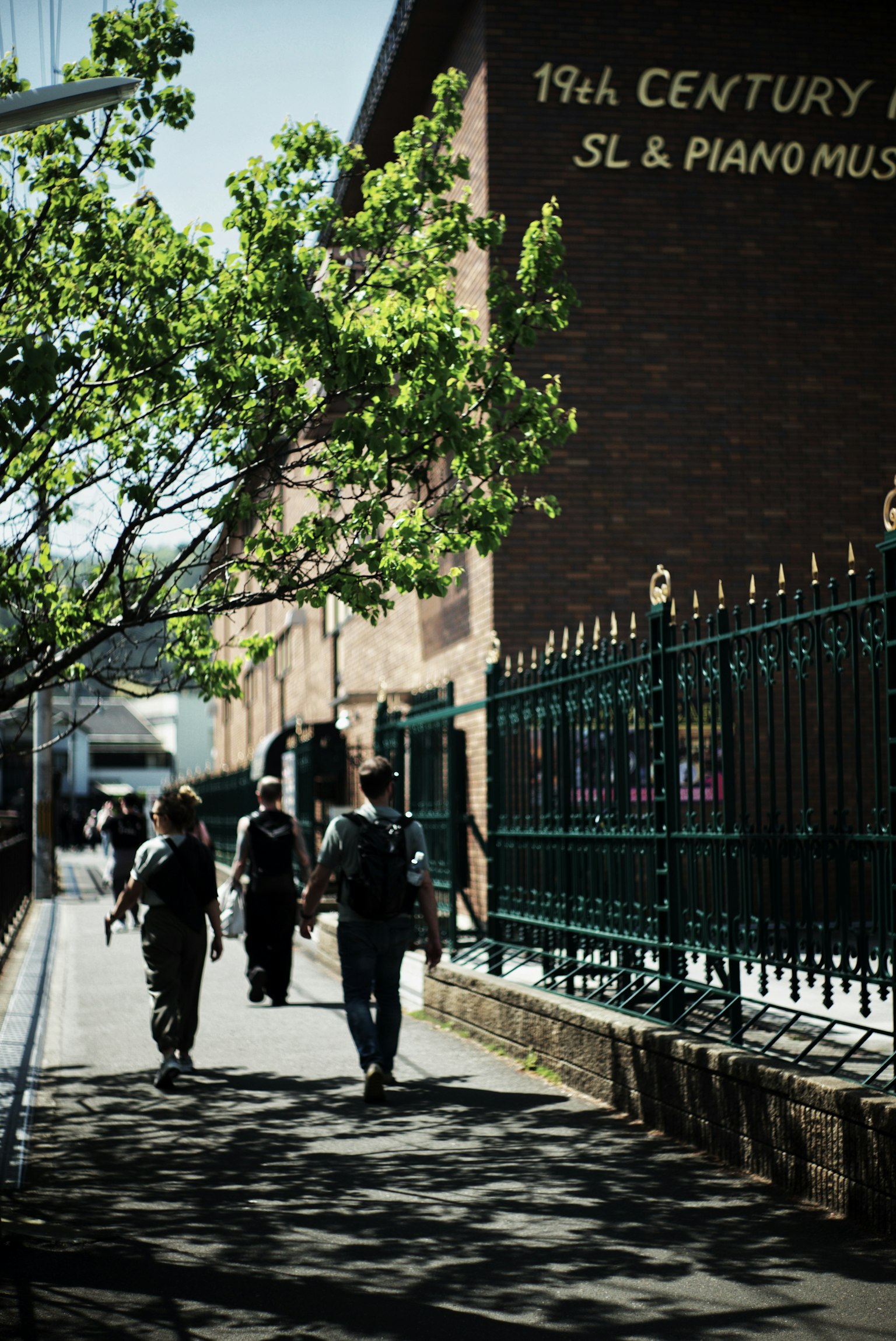 People walking along a street beside a 19th-century building with green trees and a fence