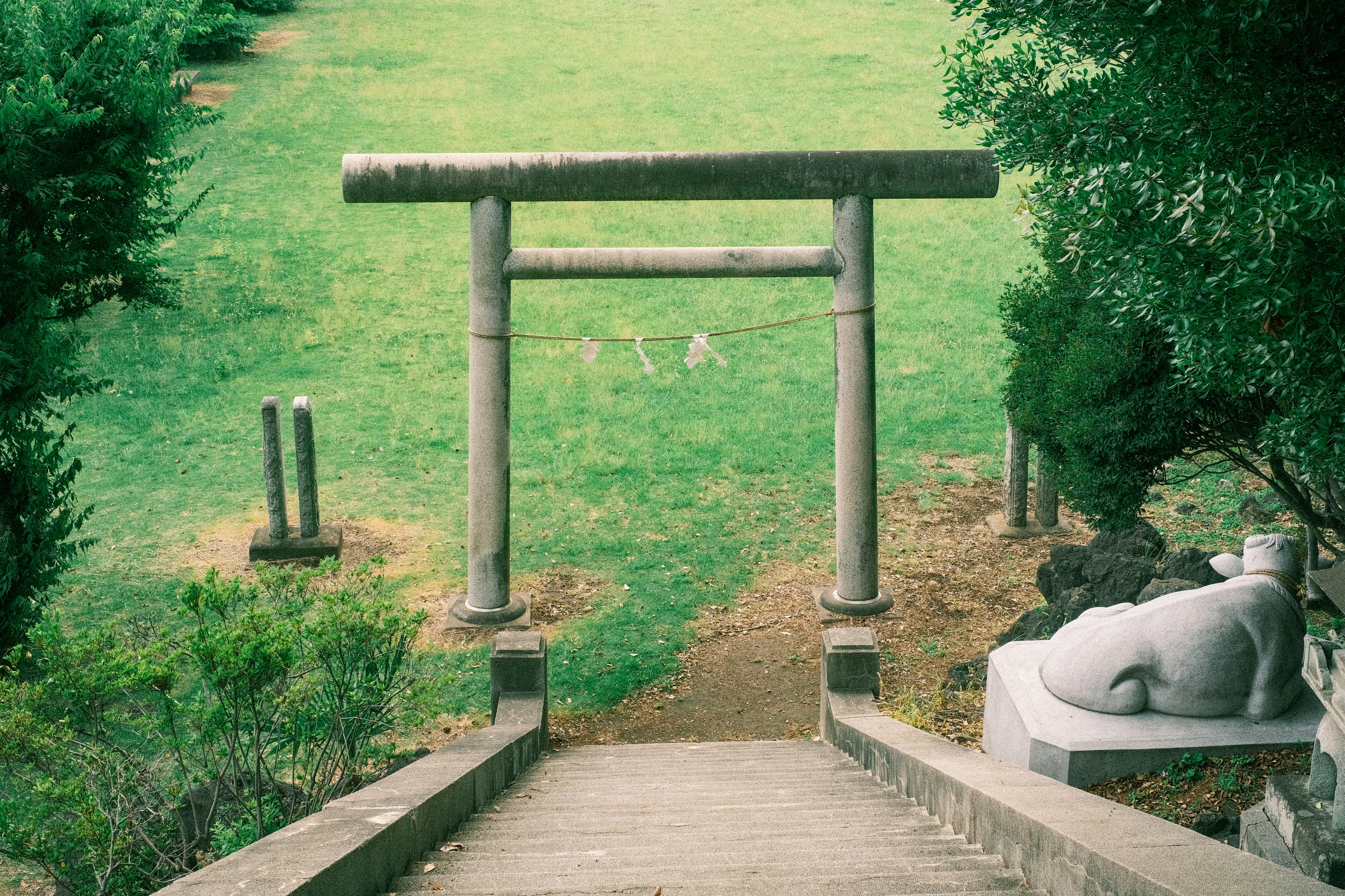Puerta torii de piedra con paisaje de césped verde y una estatua al fondo