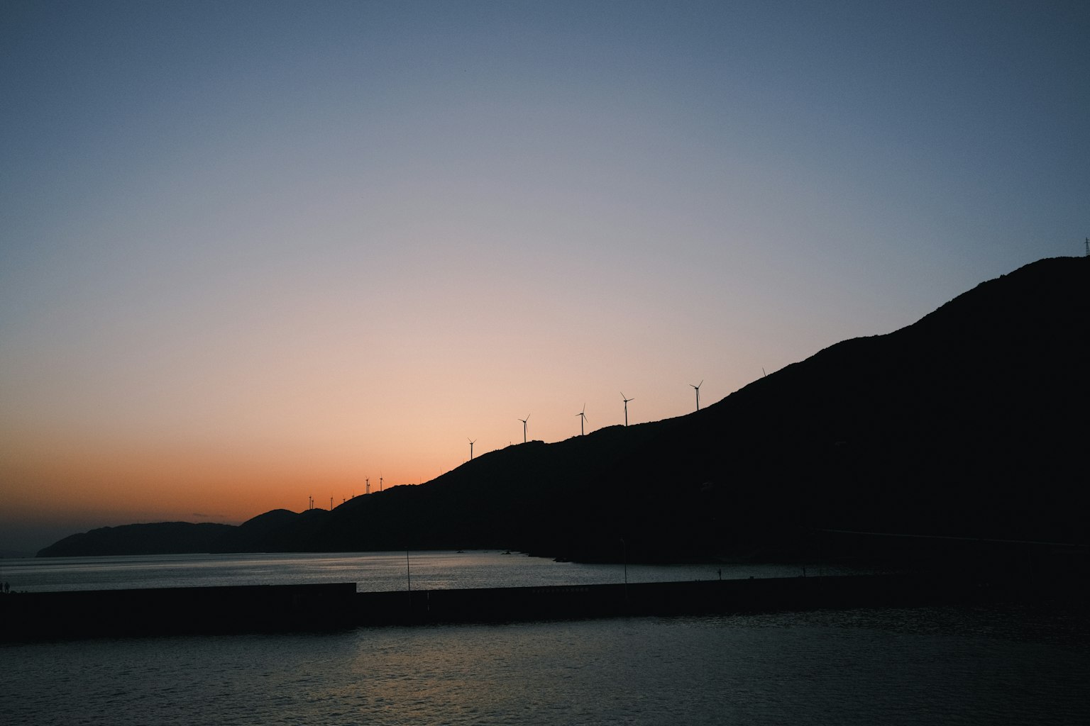 Silhouette of mountains at dusk with wind turbines visible against the sea