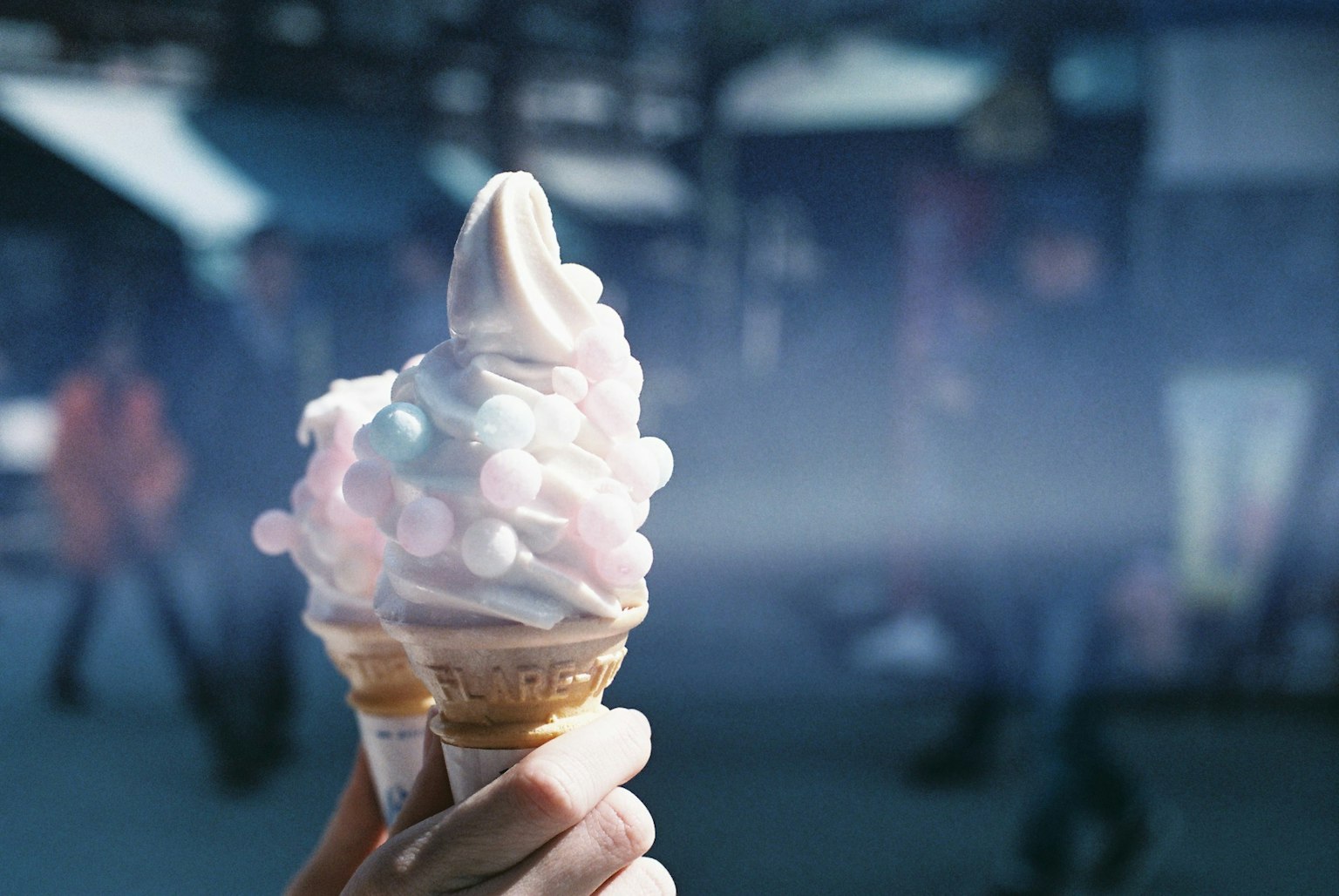 Tangan memegang cone es krim dengan es krim lembut dan taburan warna-warni di latar belakang buram orang-orang