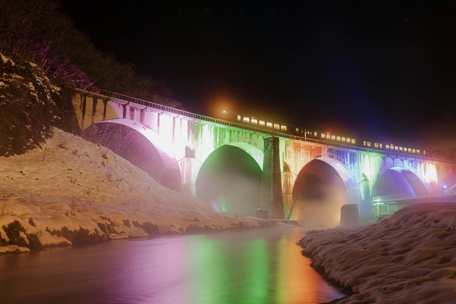 Un ponte illuminato di notte che riflette colori su un fiume innevato