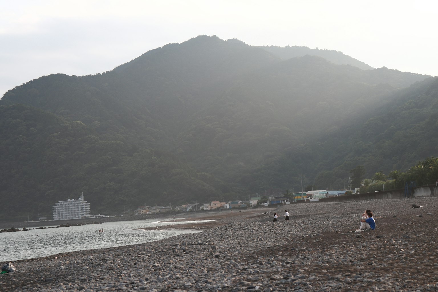 Steinstrand mit Bergen im Hintergrund und Menschen, die sitzen