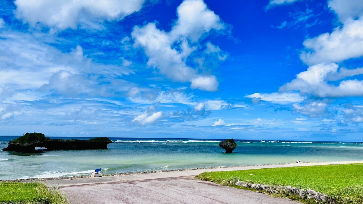 ทิวทัศน์ชายหาดที่มีท้องฟ้าสีฟ้าและเมฆสีขาวหญ้าสีเขียวและน้ำที่สงบ
