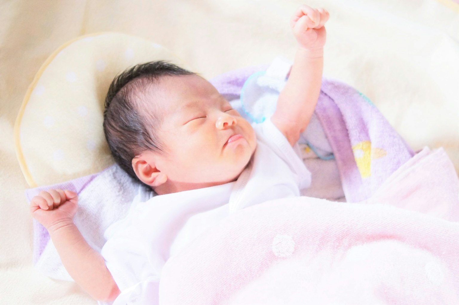 A baby sleeping with a raised arm wearing a white outfit on a soft blanket