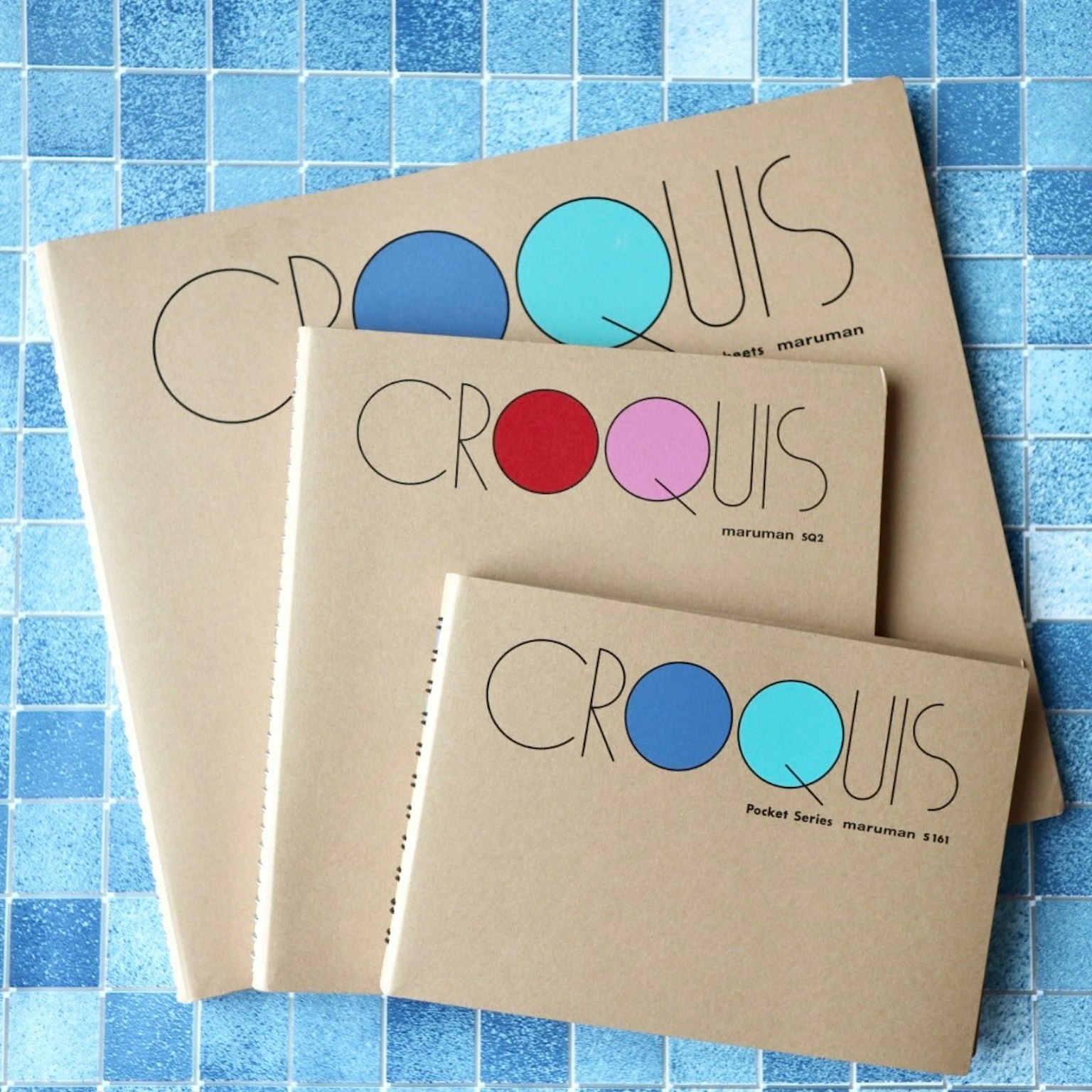 Trois carnets de croquis posés sur une surface carrelée bleue Chaque carnet présente un design de cercle coloré différent