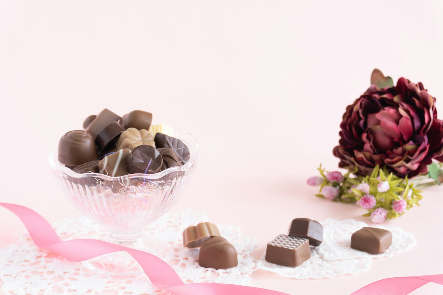 Un tazón de vidrio lleno de chocolates variados junto a una flor sobre un fondo rosa