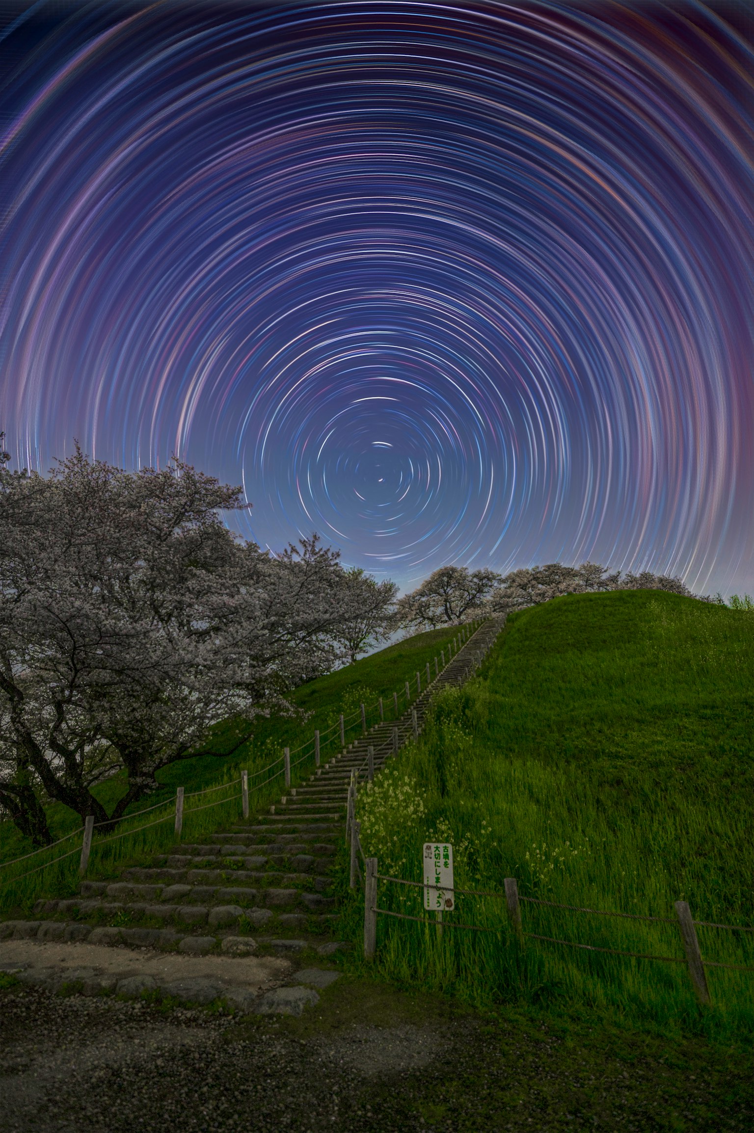 Night sky with star trails above a green hill and staircase