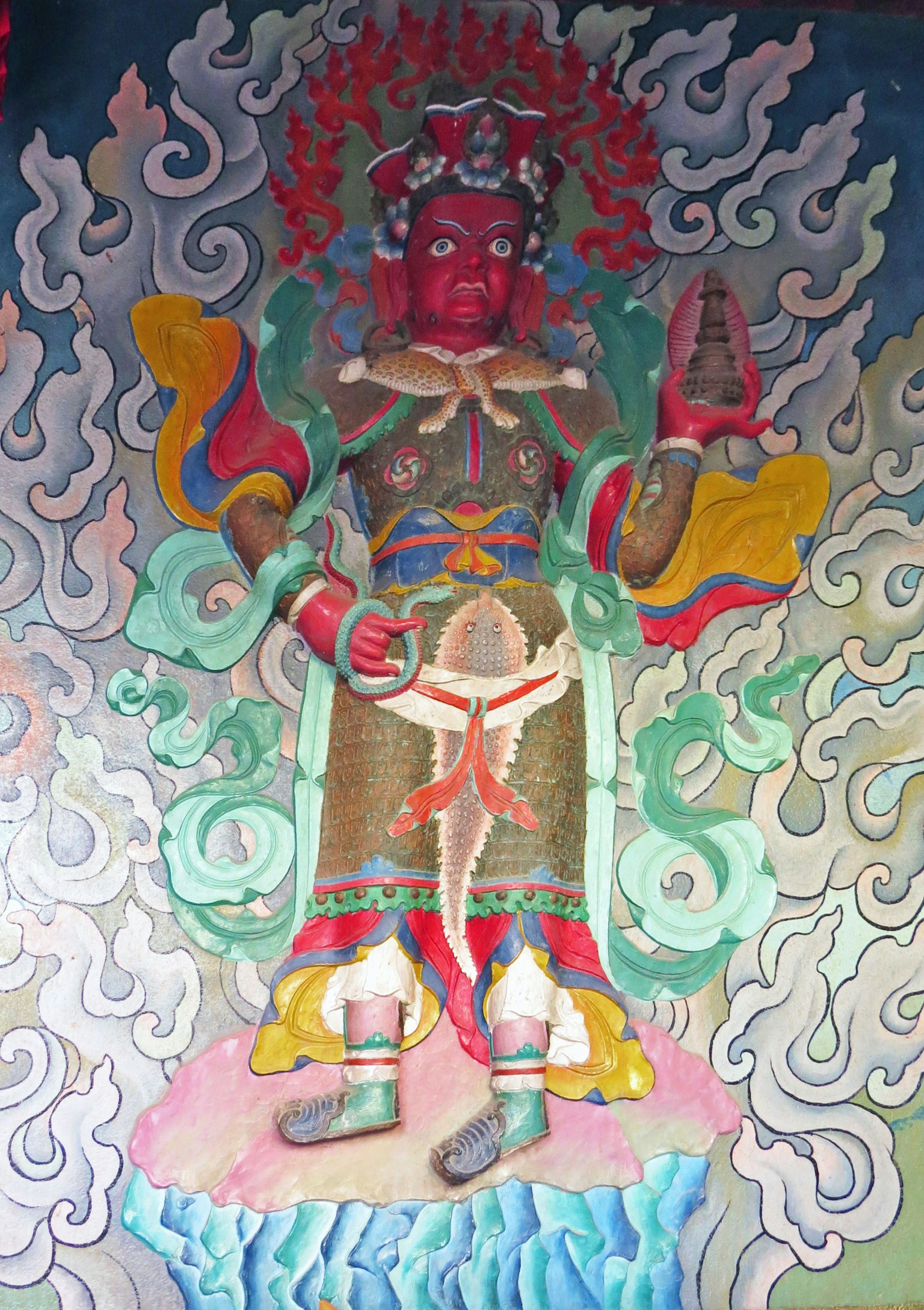A deity with a red face dressed in colorful attire surrounded by cloud patterns in the background